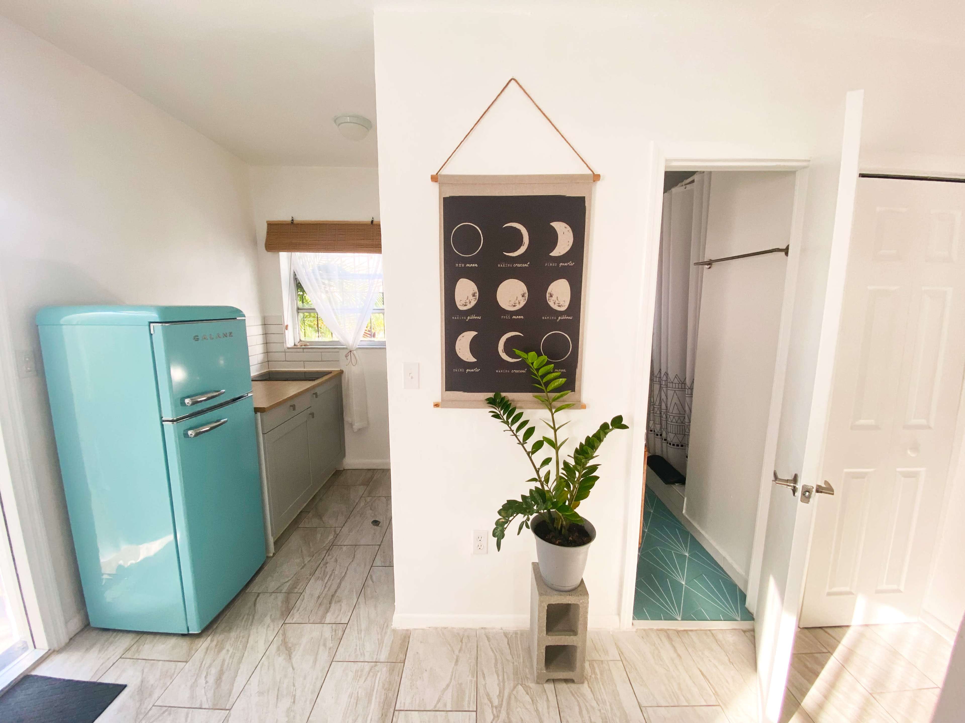The image shows a bright, minimalist kitchen area featuring a turquoise refrigerator, a plant in a pot, and a wall hanging displaying phases of the moon.