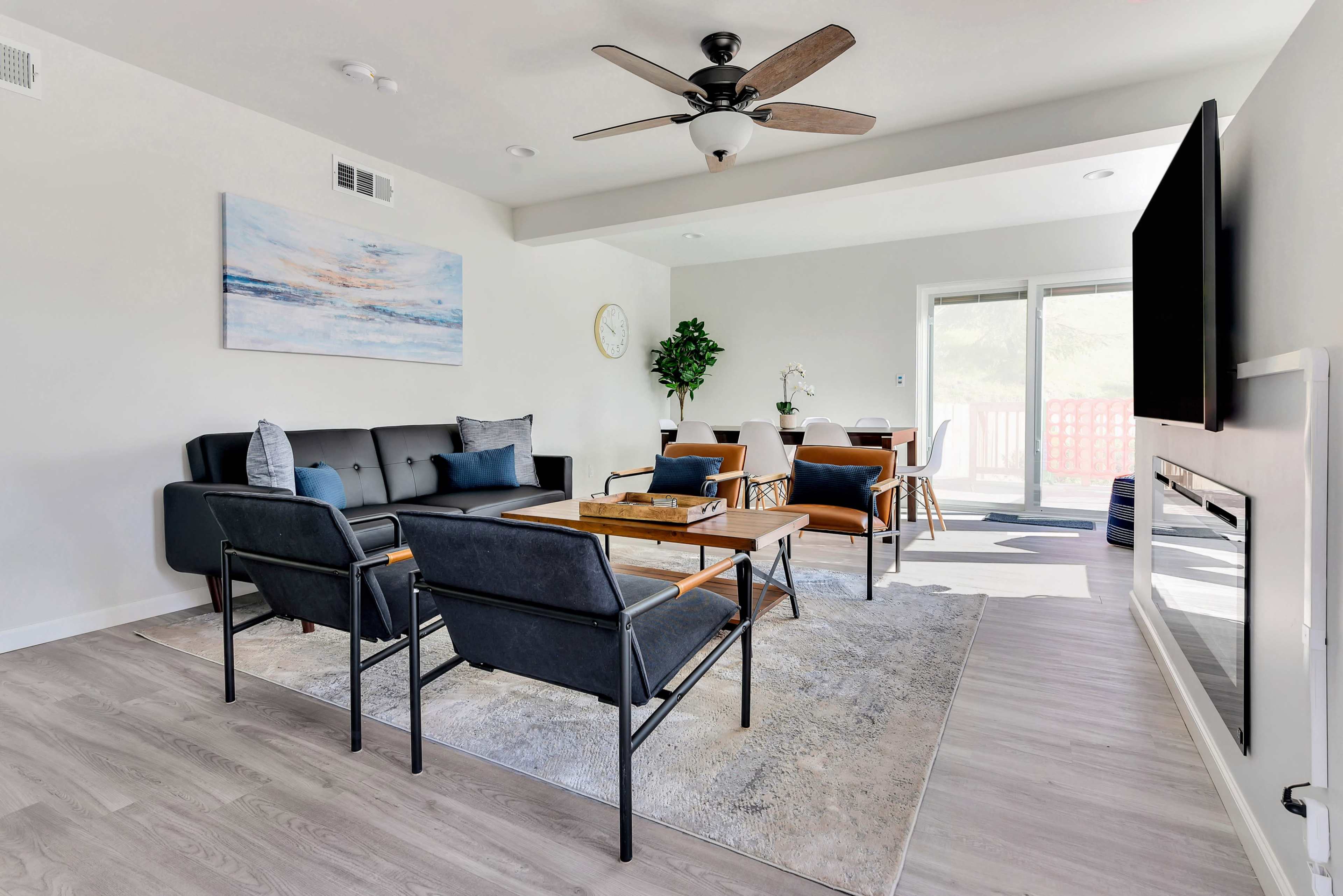 A modern living room features a black sofa, two chairs, a coffee table, and a wall-mounted television, with large windows allowing natural light.
