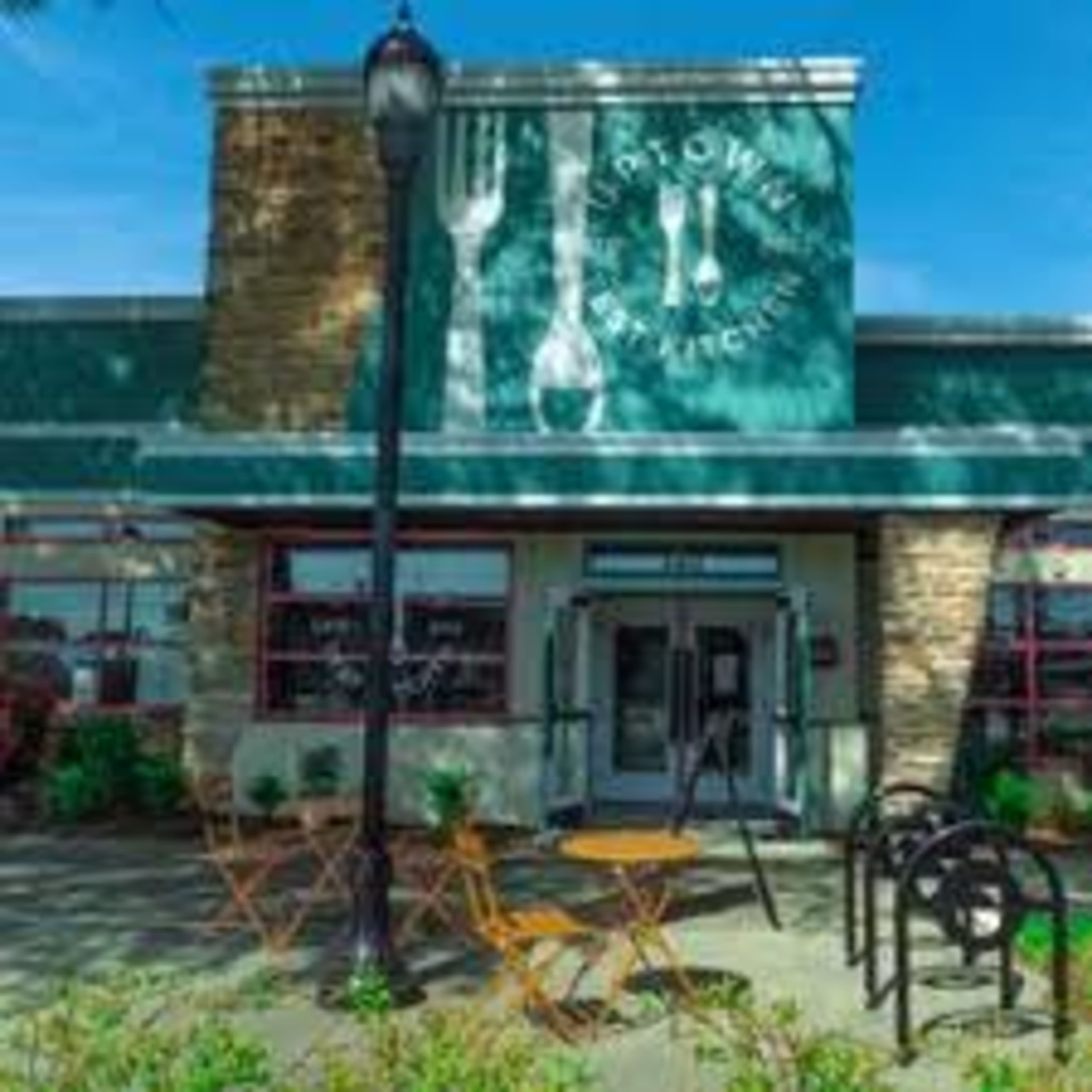 The image shows the exterior of a restaurant with a large sign featuring utensils on the wall and outdoor seating in front.