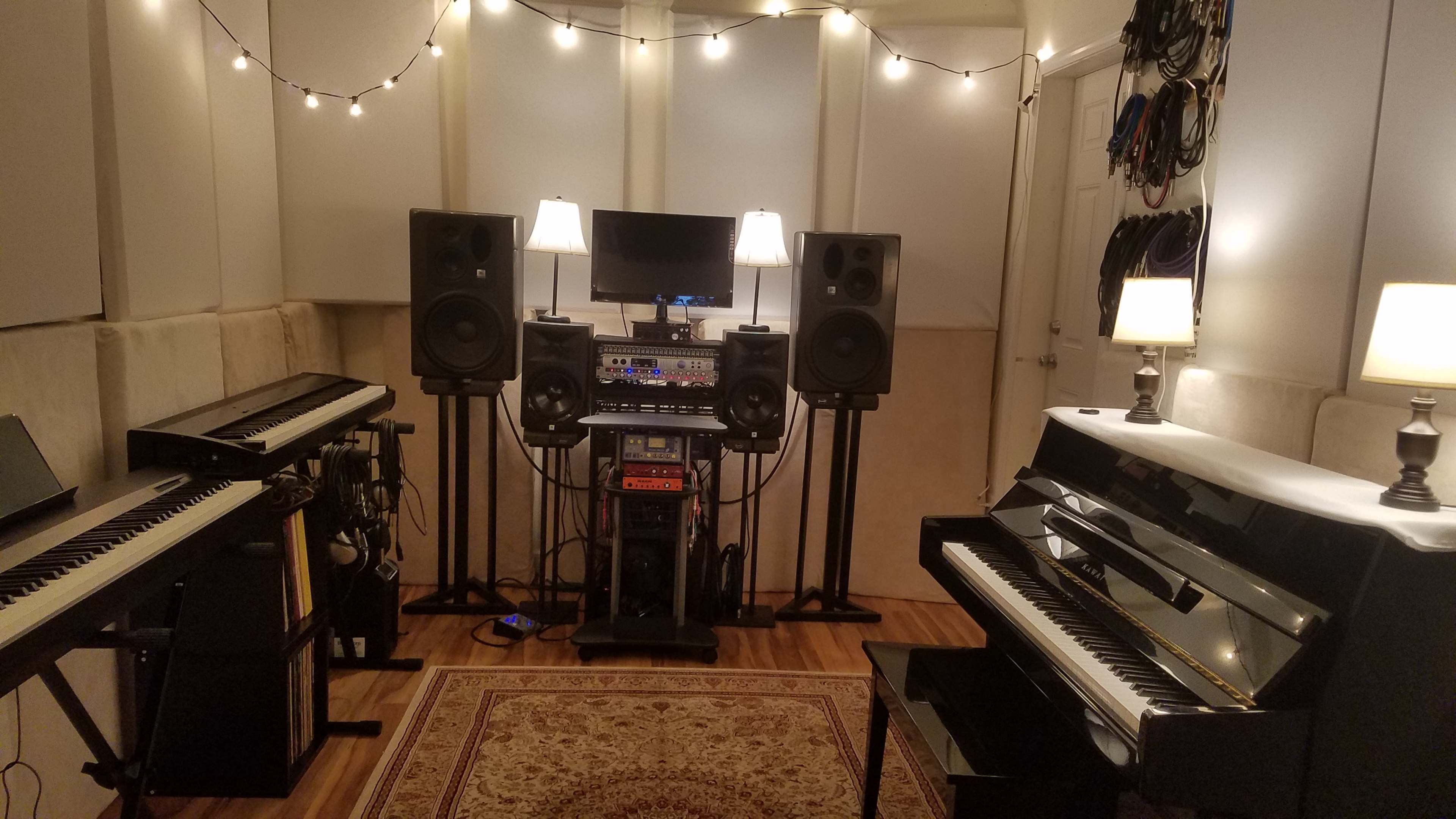 A music studio with two keyboards on either side, a rack of audio equipment in the center, and speakers flanking a monitor, all under warm lighting from lamps.