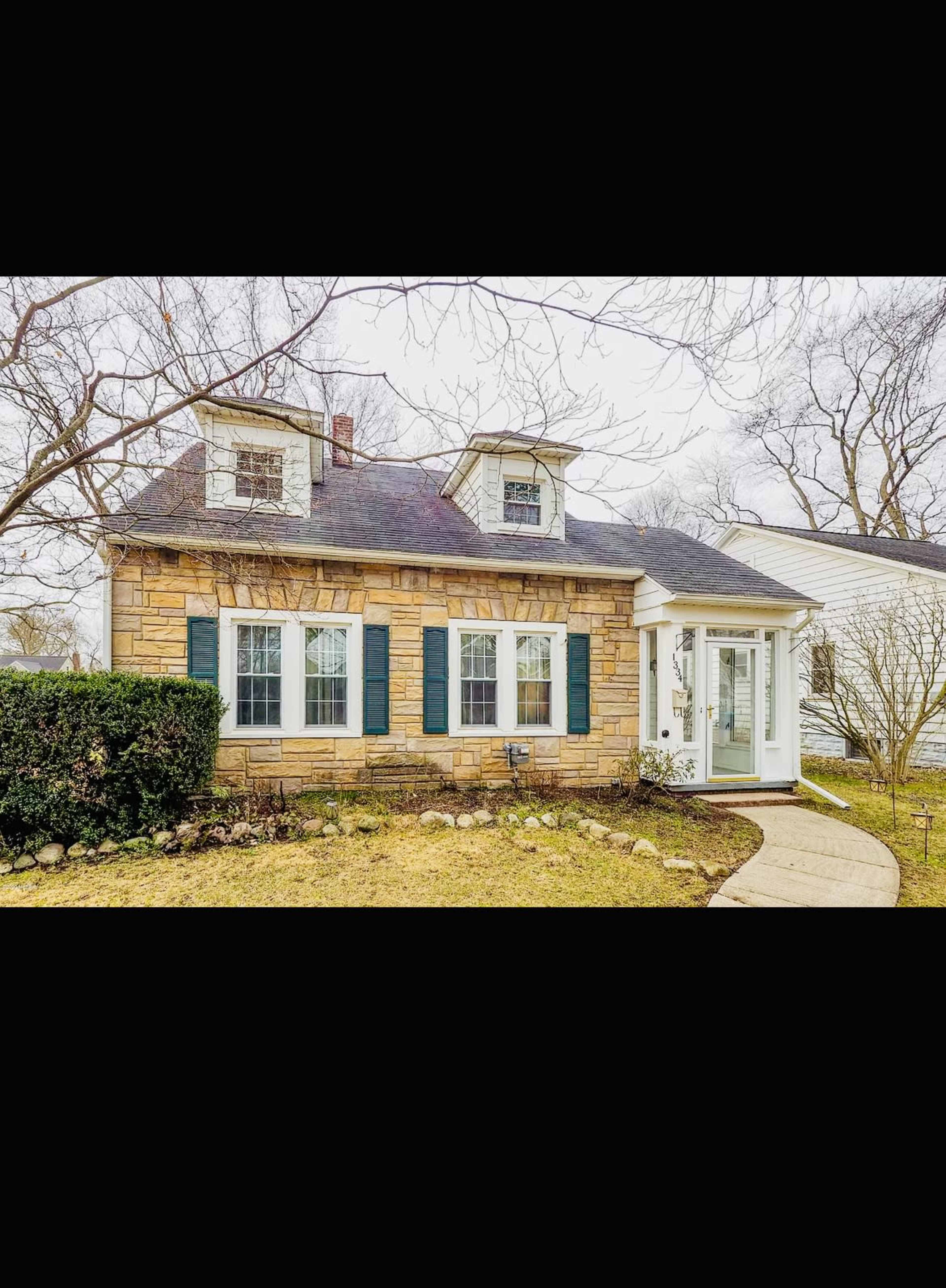 A stone-clad house with a peaked roof, dark shutters, and a front porch surrounded by a manicured lawn and shrubs.