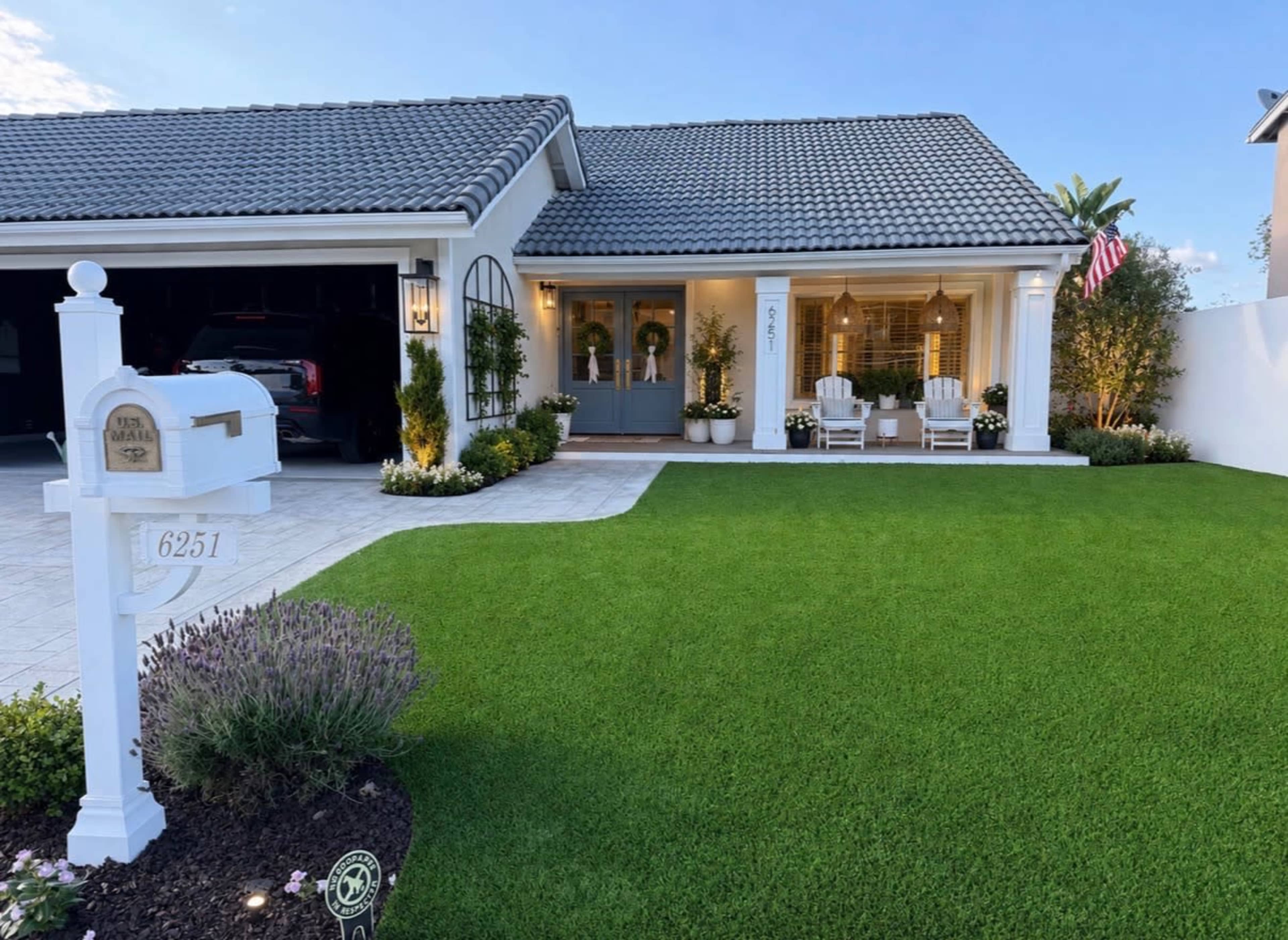 A well-manicured front yard features a decorative mailbox, a driveway leading to a garage, and a house with a tiled roof and a porch adorned with rocking chairs and potted plants.