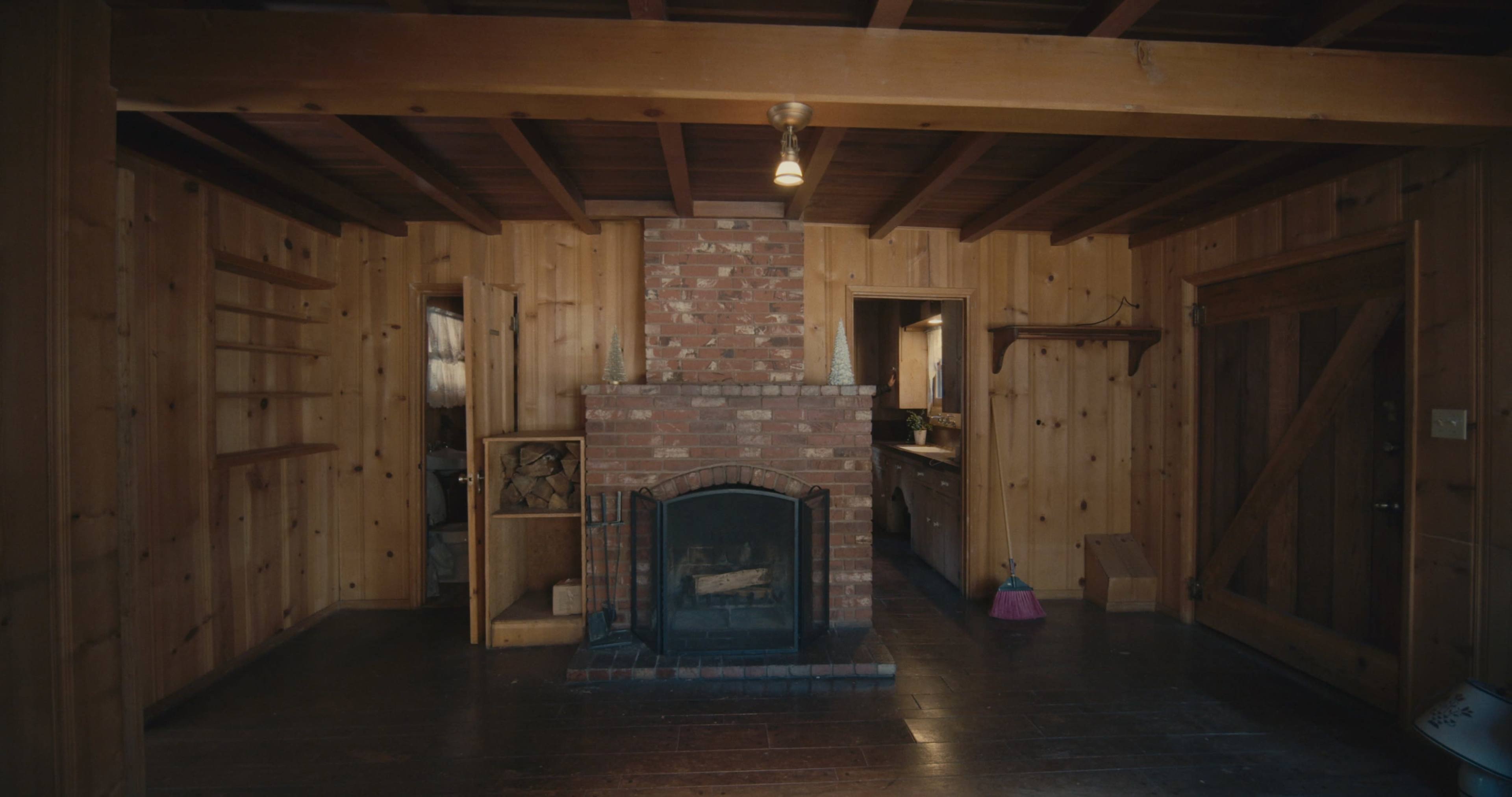 The image shows a wooden-paneled room with a brick fireplace centered against one wall, leading to a doorway that opens into a kitchen area.