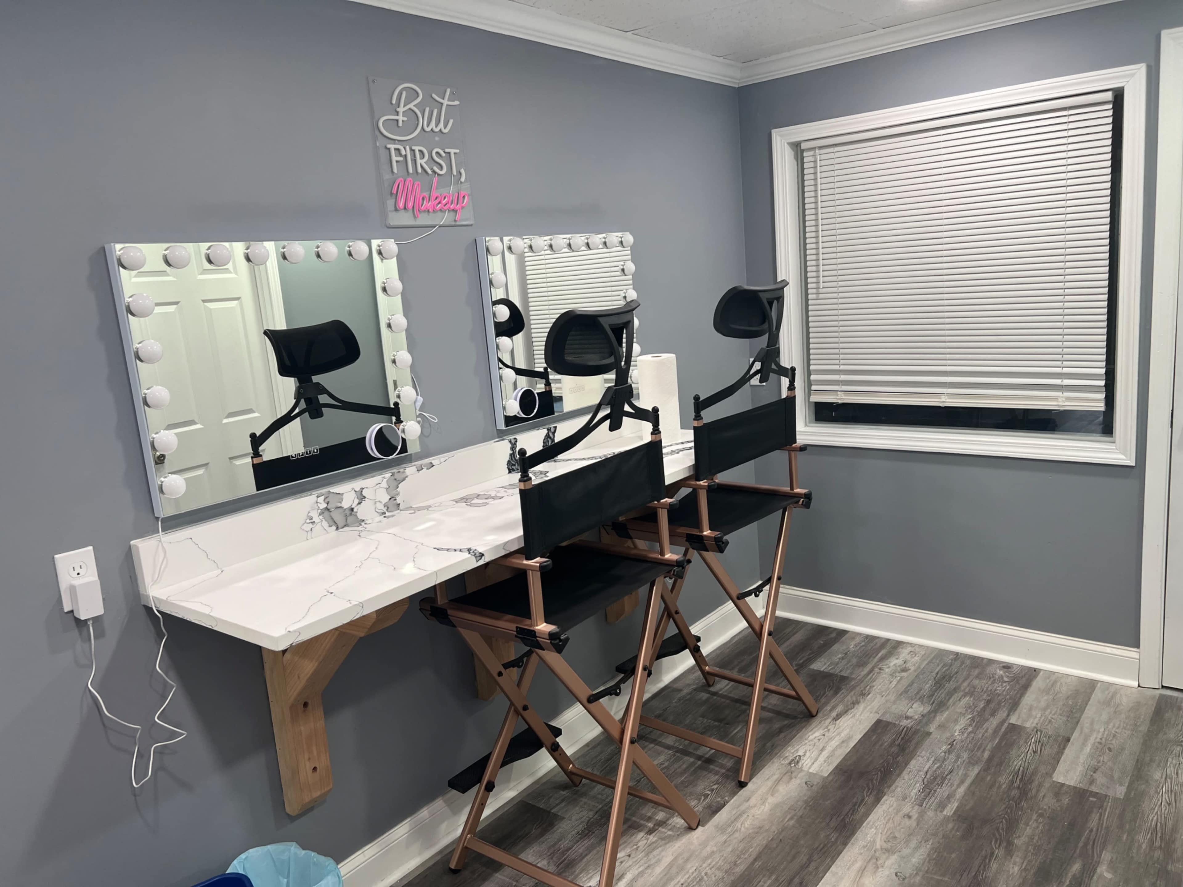 The image shows a makeup station featuring two director's chairs, a large illuminated mirror, and a window with closed blinds against a gray wall.