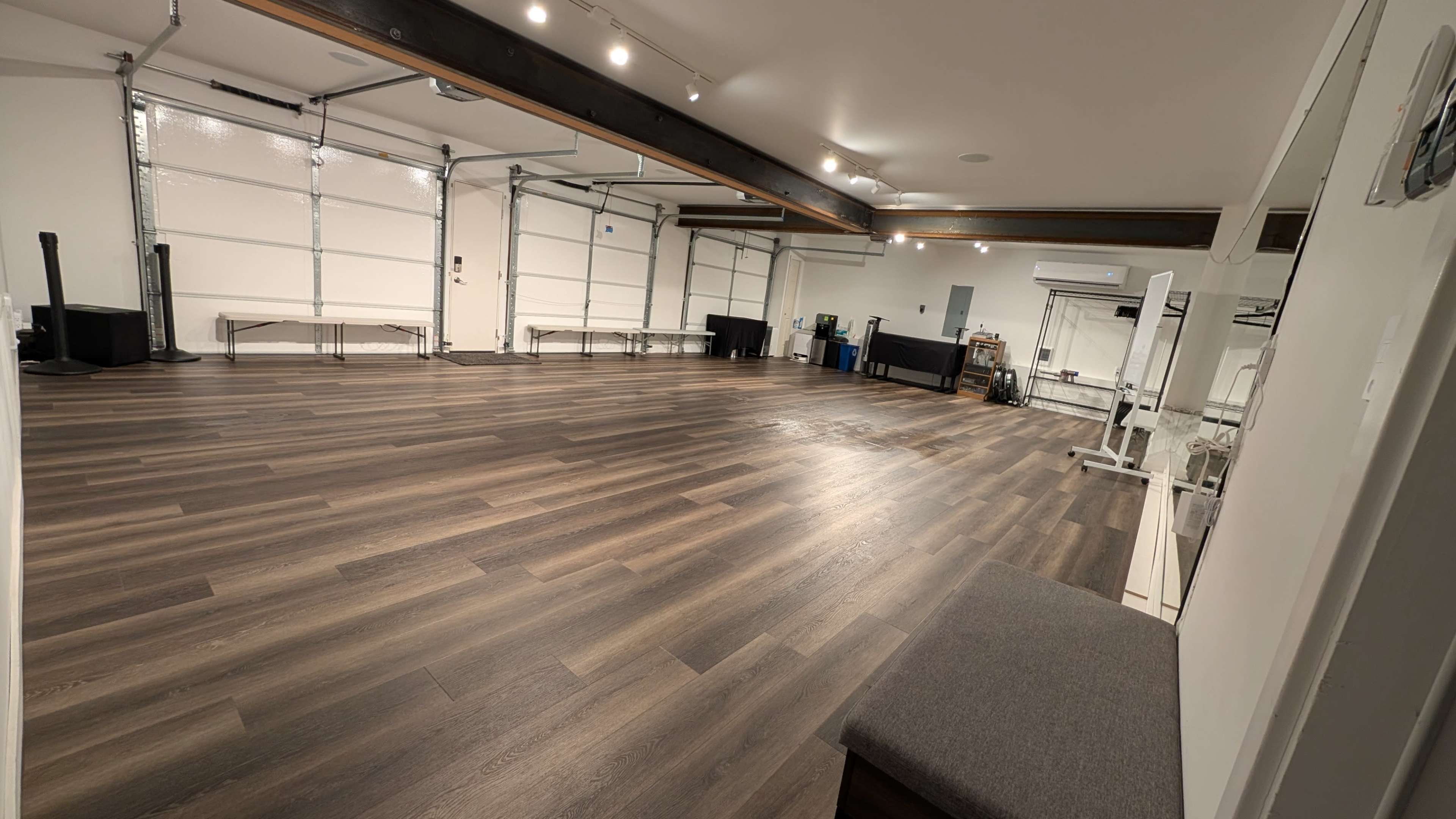 The image shows a spacious interior room with a polished wooden floor, equipped with workout equipment and two large garage doors.