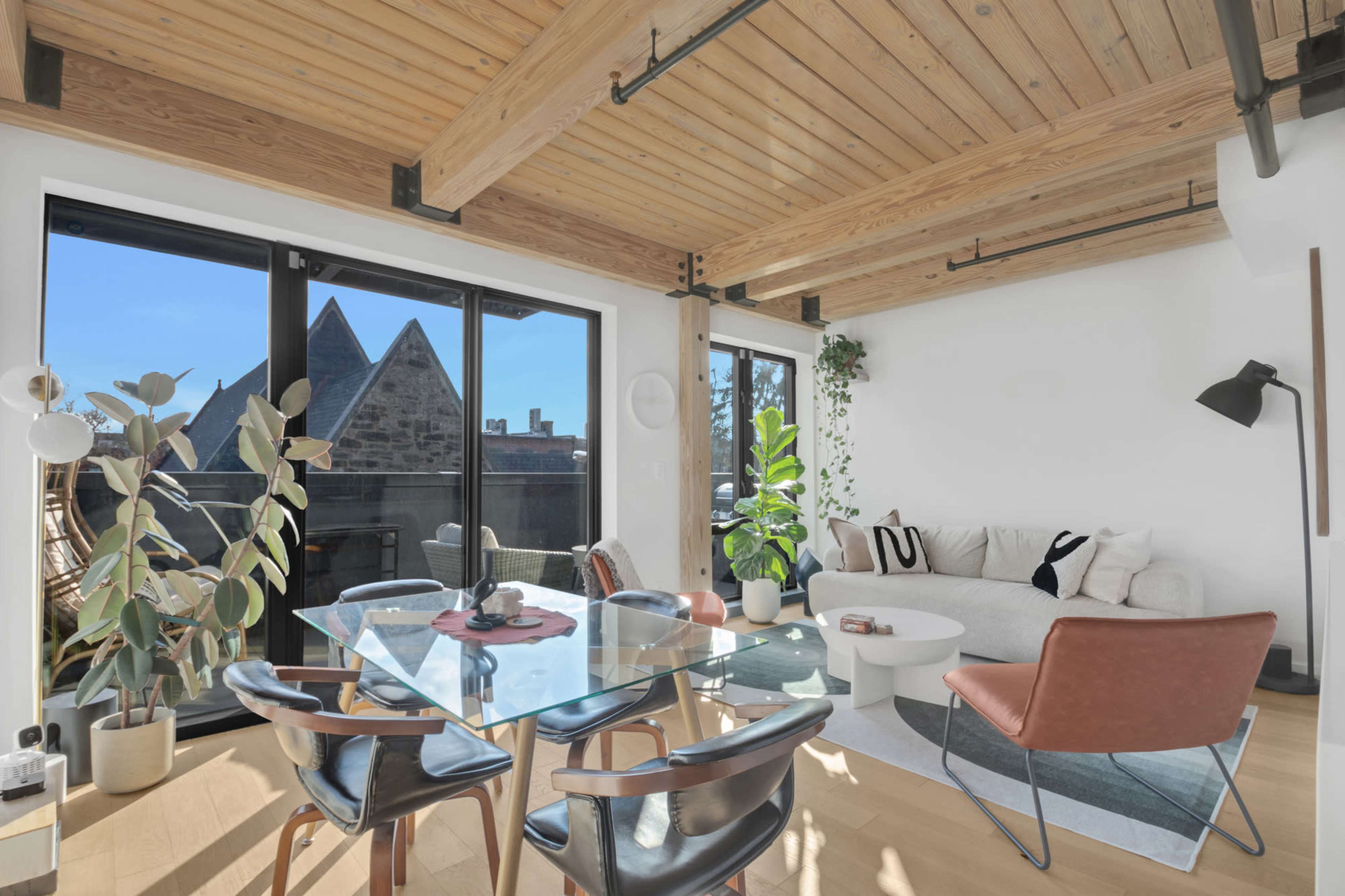 The image shows a modern living space with large windows, wooden beams, a glass dining table, and contemporary furniture.