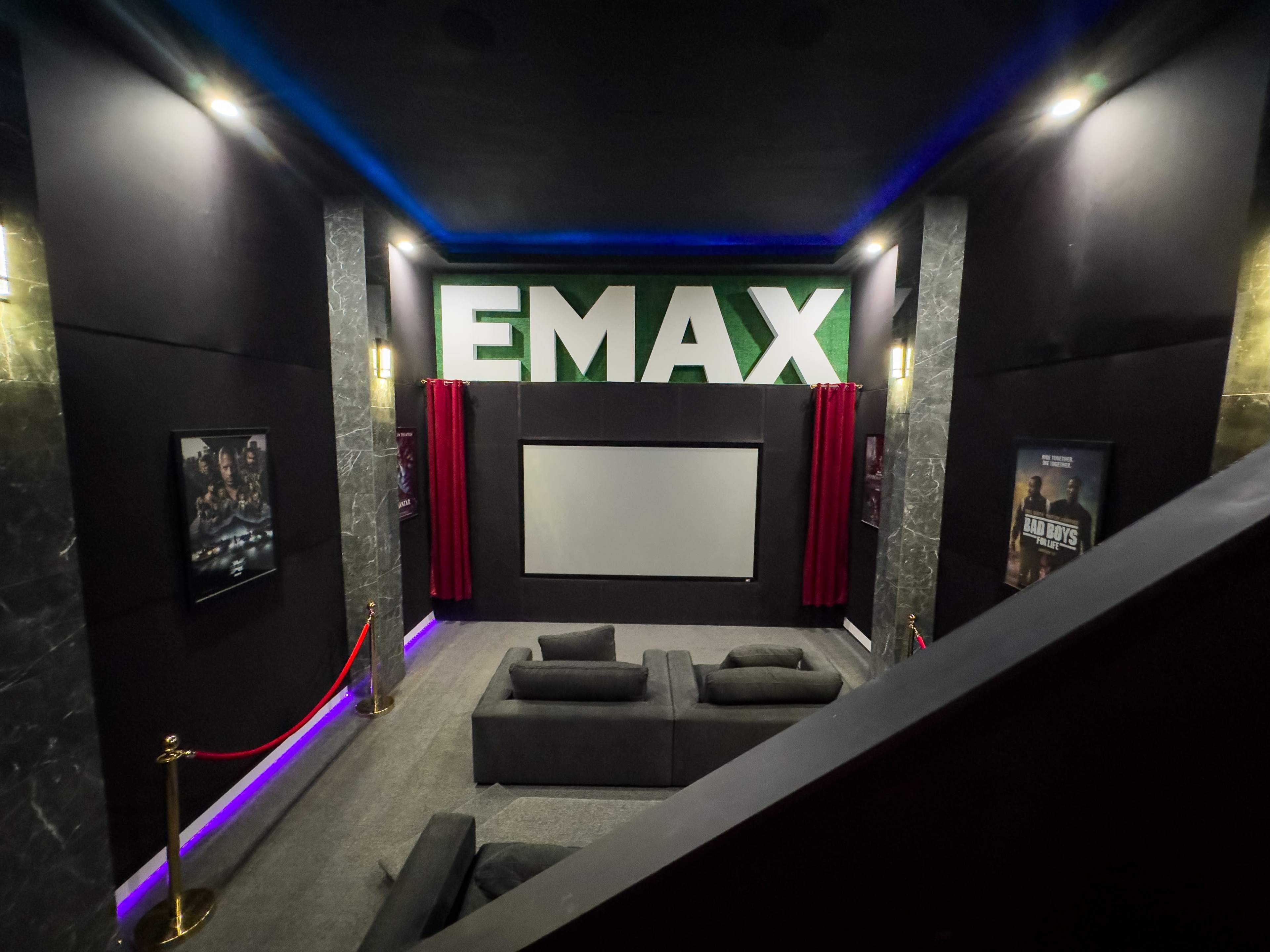 The image shows a modern home theater with large "EMAX" signage, a projector screen, plush seating, and decorative lighting.