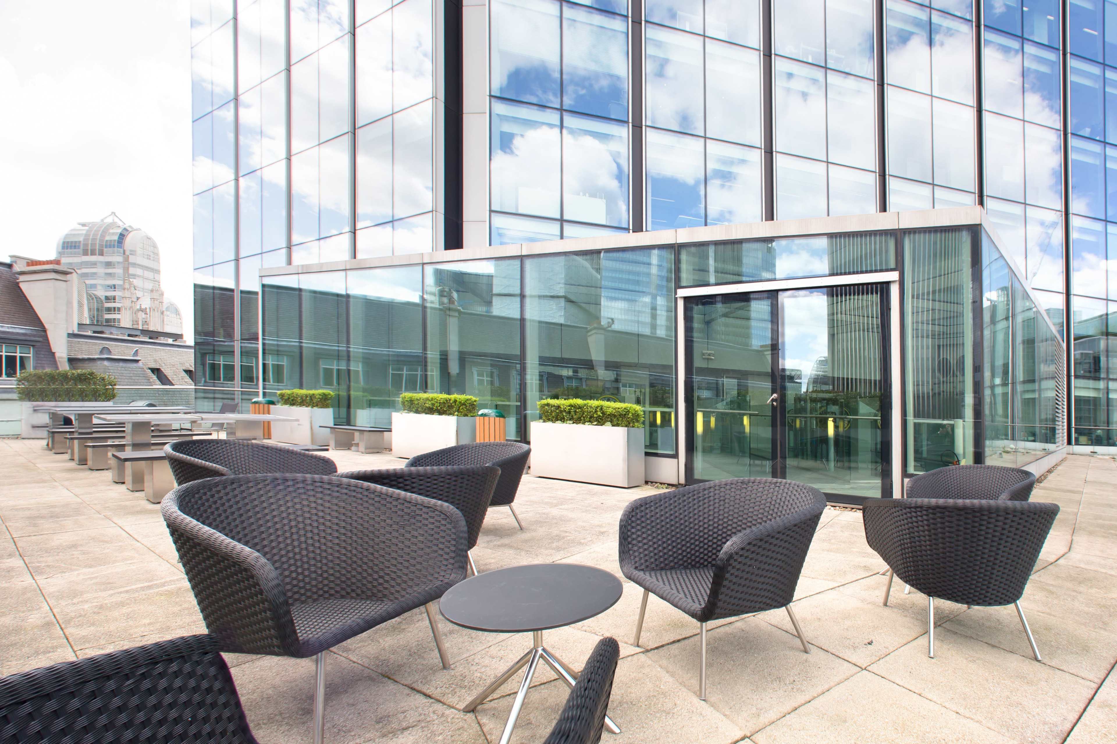 A rooftop terrace features modern furniture and large glass windows reflecting the surrounding buildings and sky.