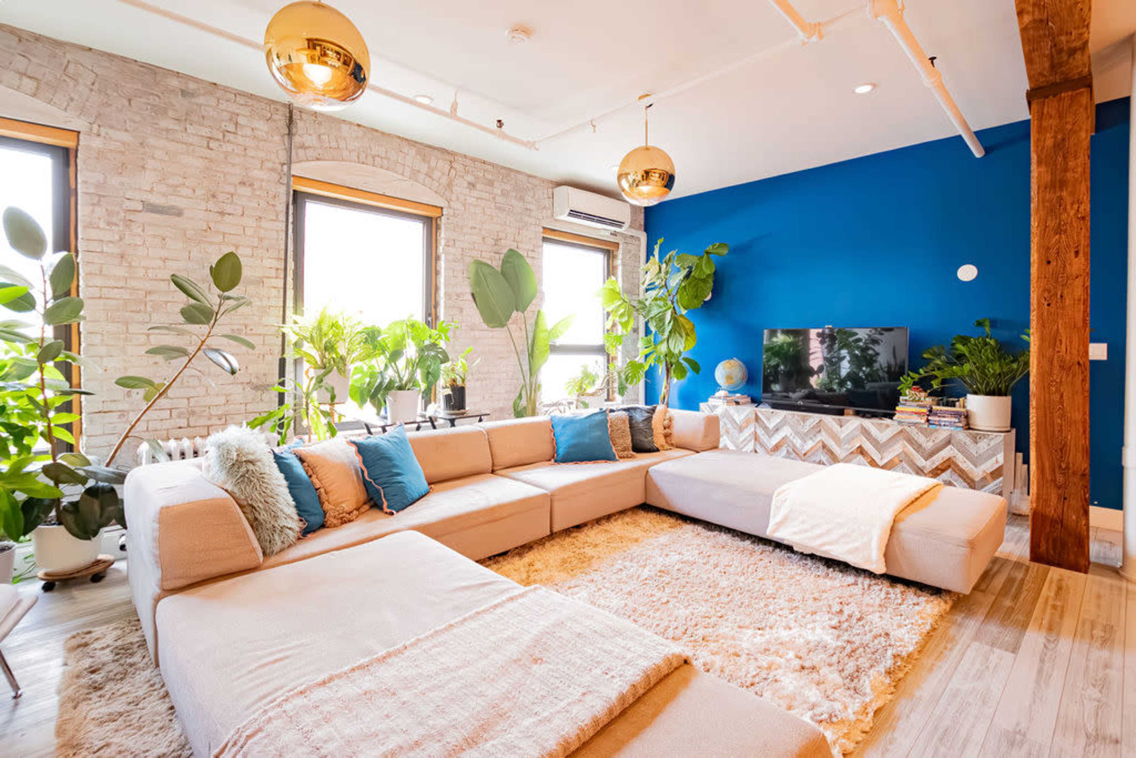 The living room features a large sectional sofa, vibrant blue accent wall, and several potted plants that create a bright and inviting atmosphere.