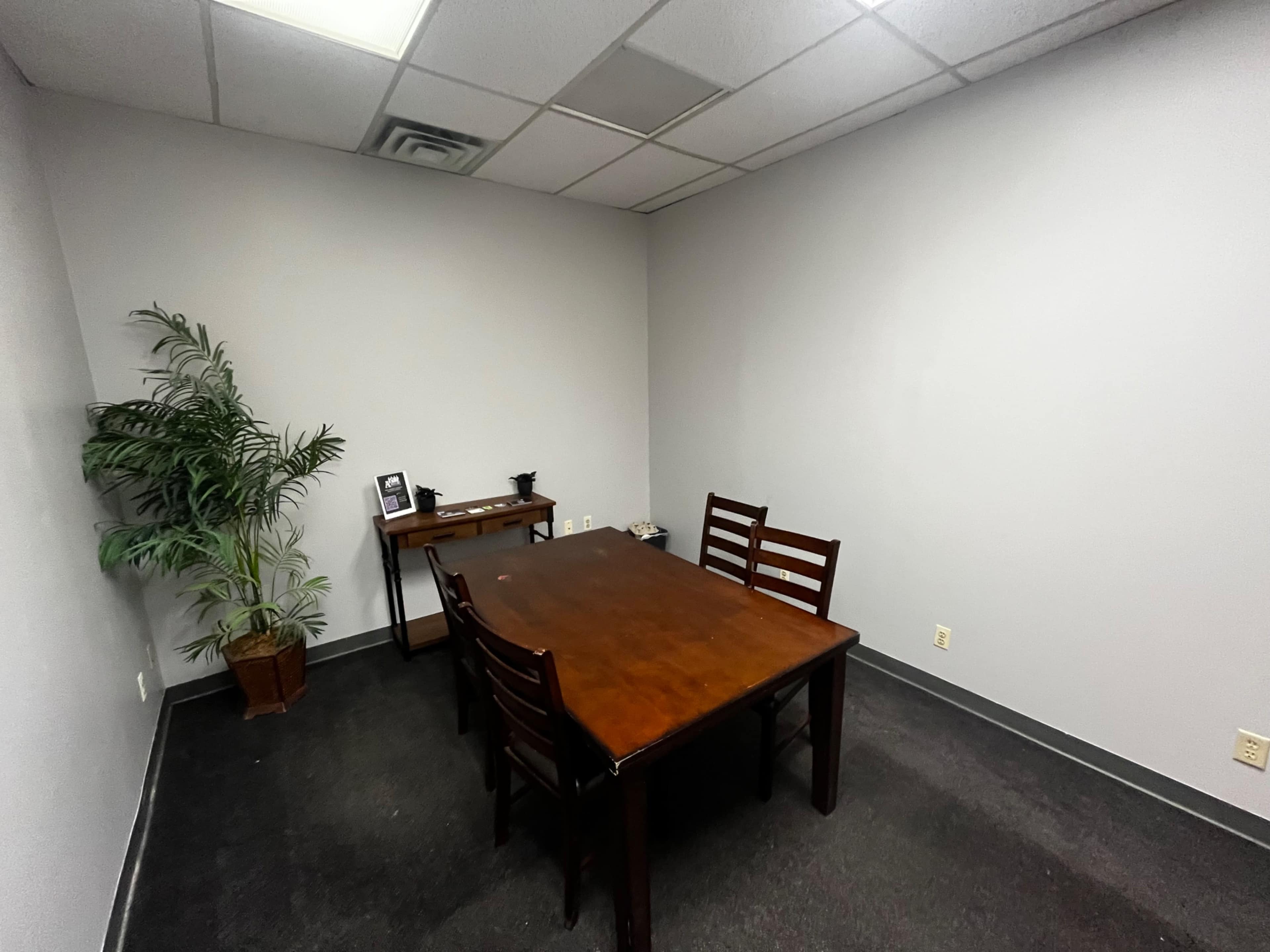 A sparsely furnished office space features a wooden table surrounded by four chairs, with a potted plant in one corner.