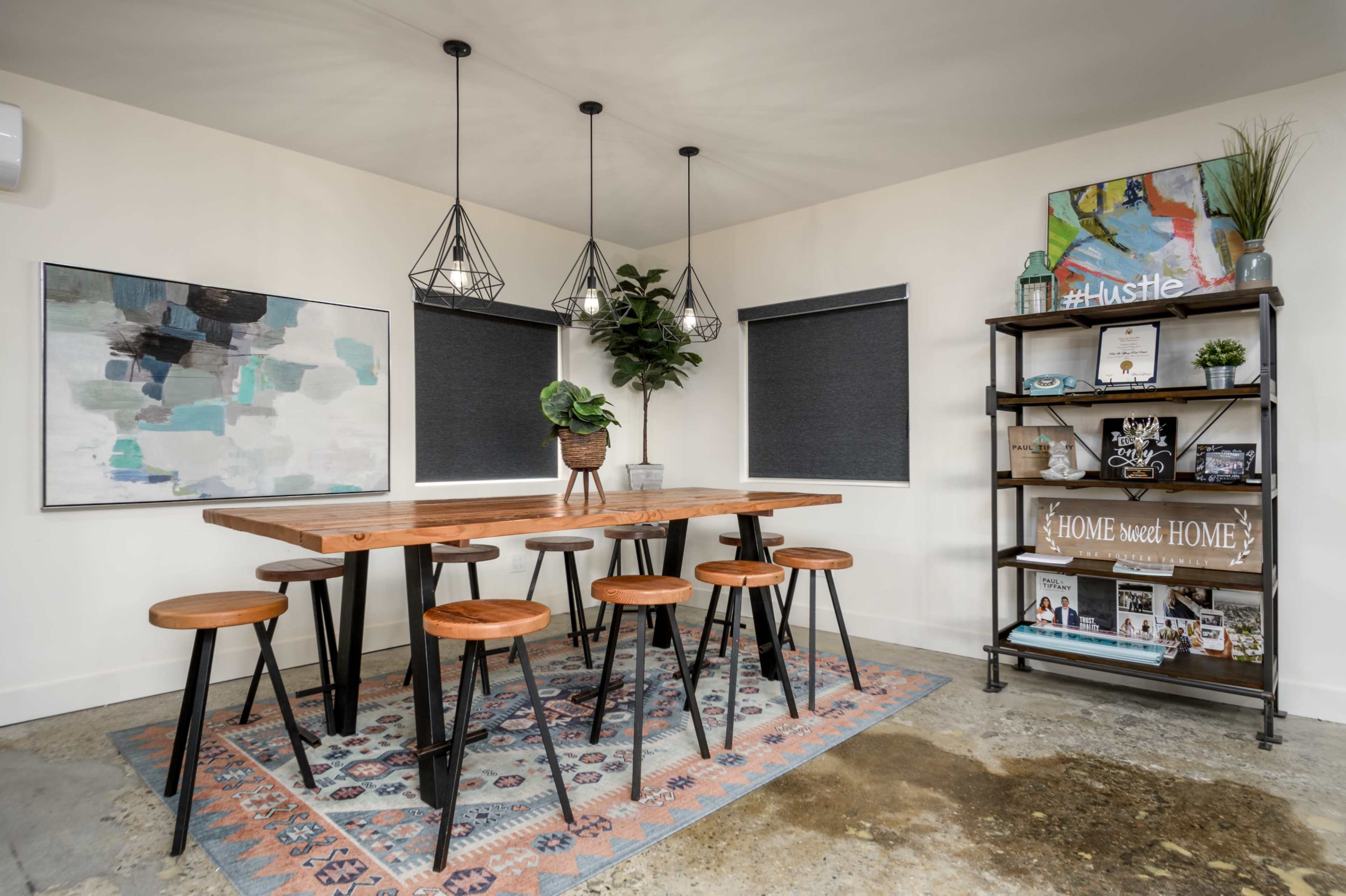 A modern meeting space with a large wooden table surrounded by high stools, decorative plants, and a bookshelf displaying various items and books.