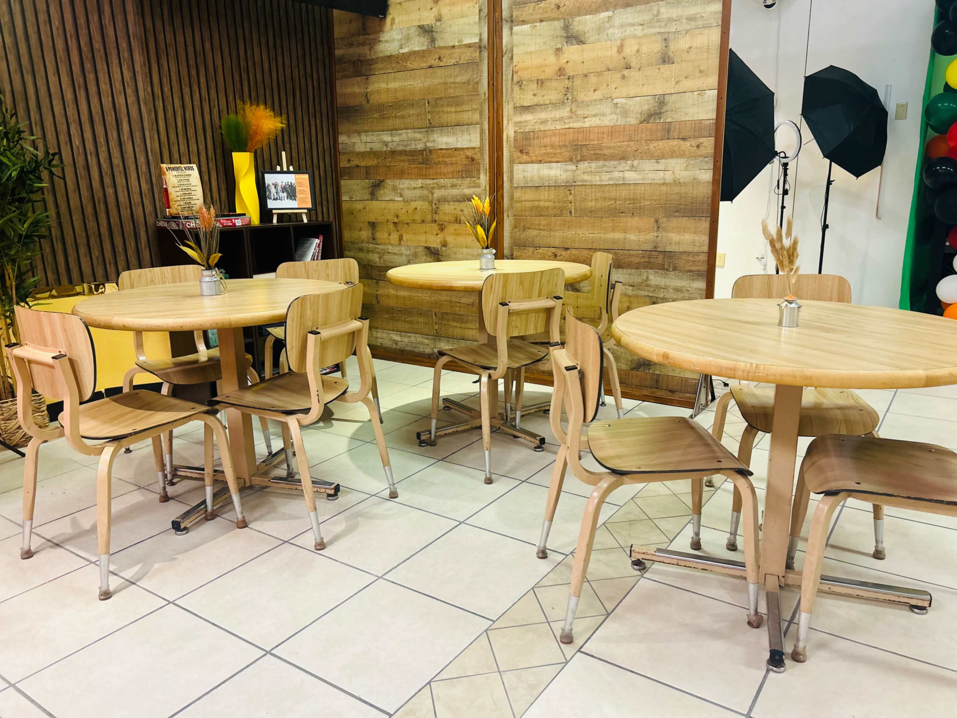 A cozy café setting with wooden tables and chairs arranged on a tiled floor, surrounded by wooden wall panels and decorative plants.