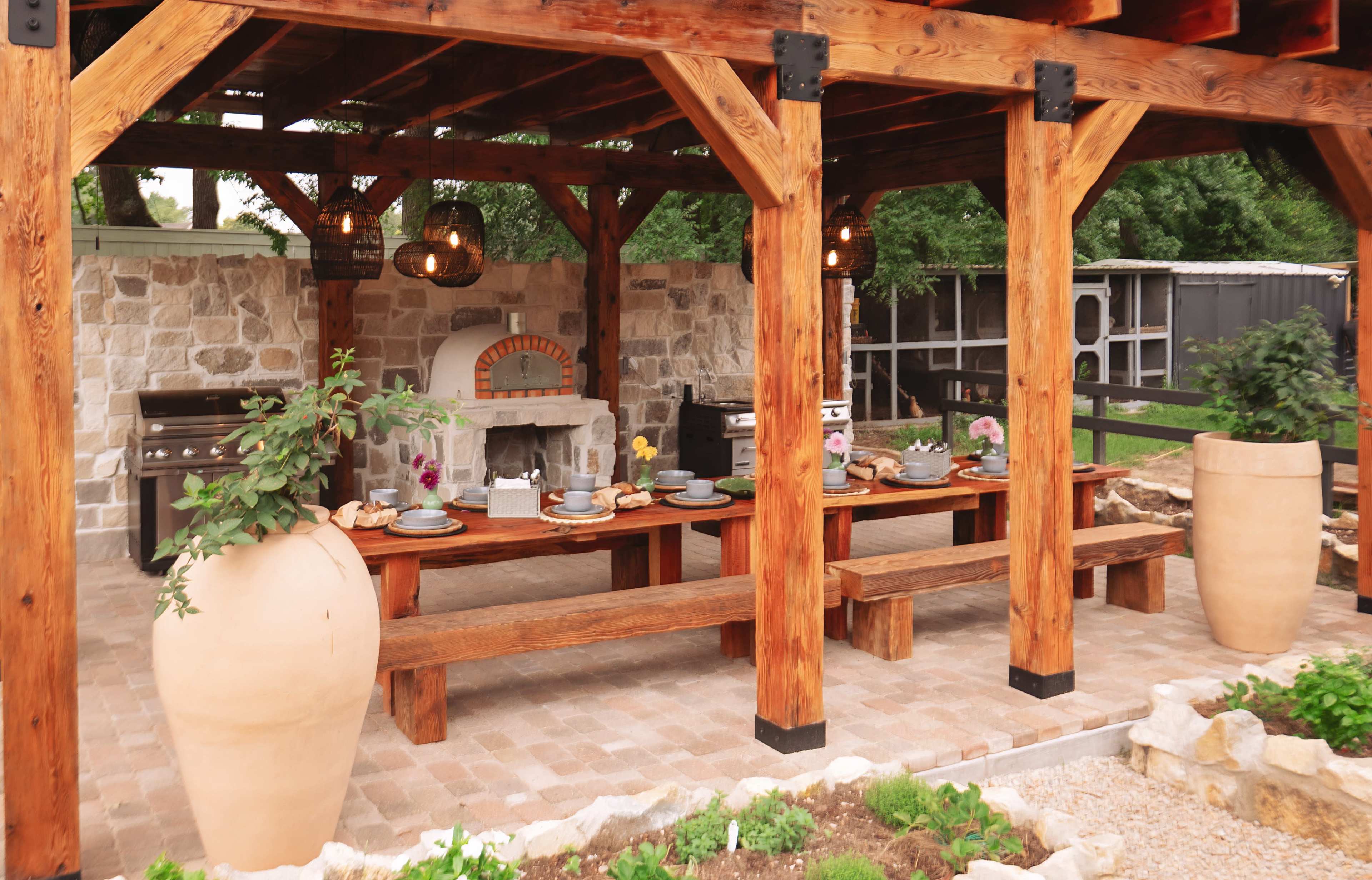 The image shows an outdoor kitchen area with a wooden pavilion, stone walls, a large dining table set with dishes, and two large ceramic vases.