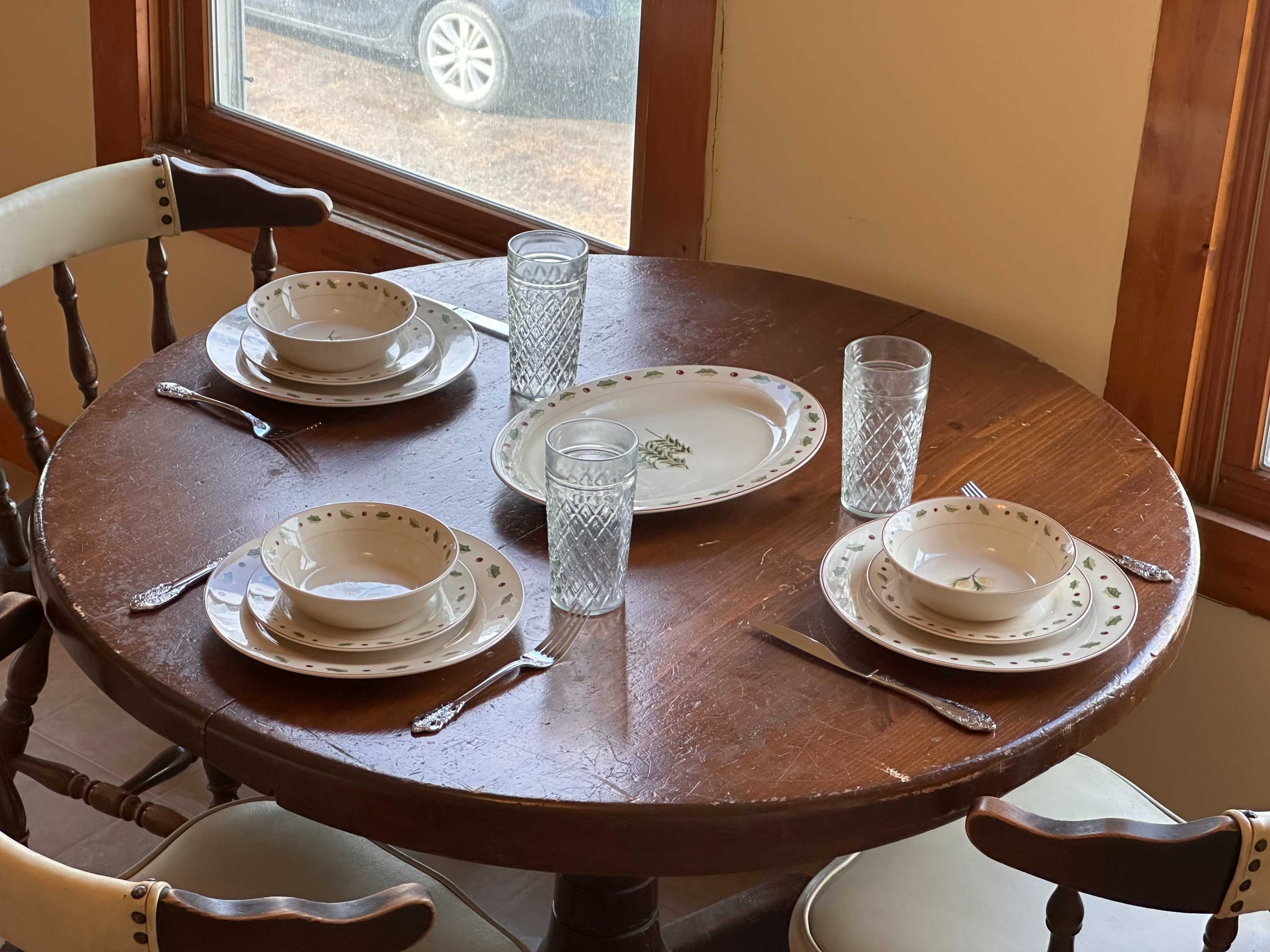 A round wooden table is set for four with plates, bowls, glasses, and utensils, beside a window showing a parked car outside.
