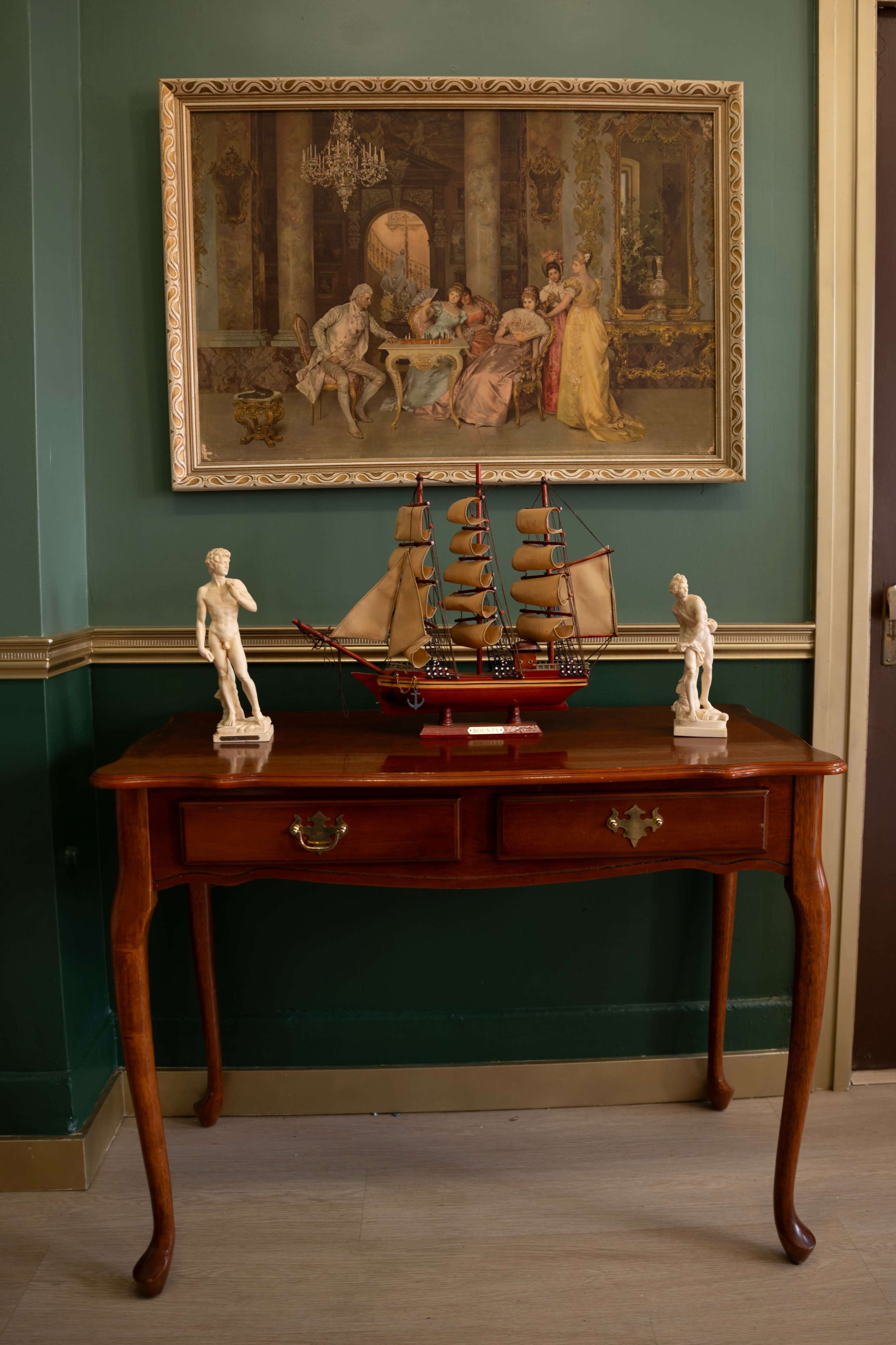 A wooden table with a ship model and two statues is set against a green wall adorned with a framed painting depicting a historical scene.