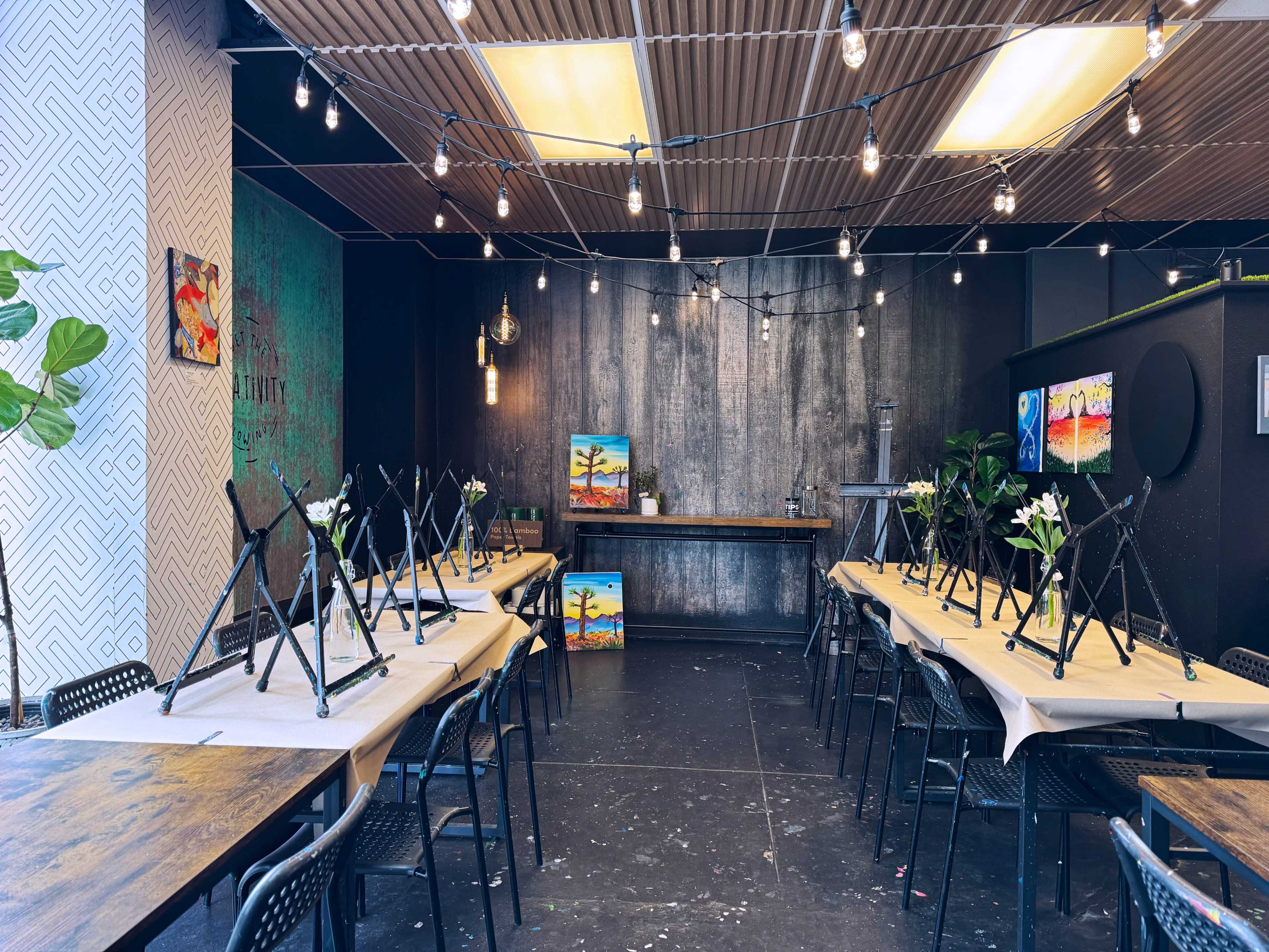 The image shows an art studio with multiple tables set up for a painting class, featuring easels, paint supplies, and string lights overhead.