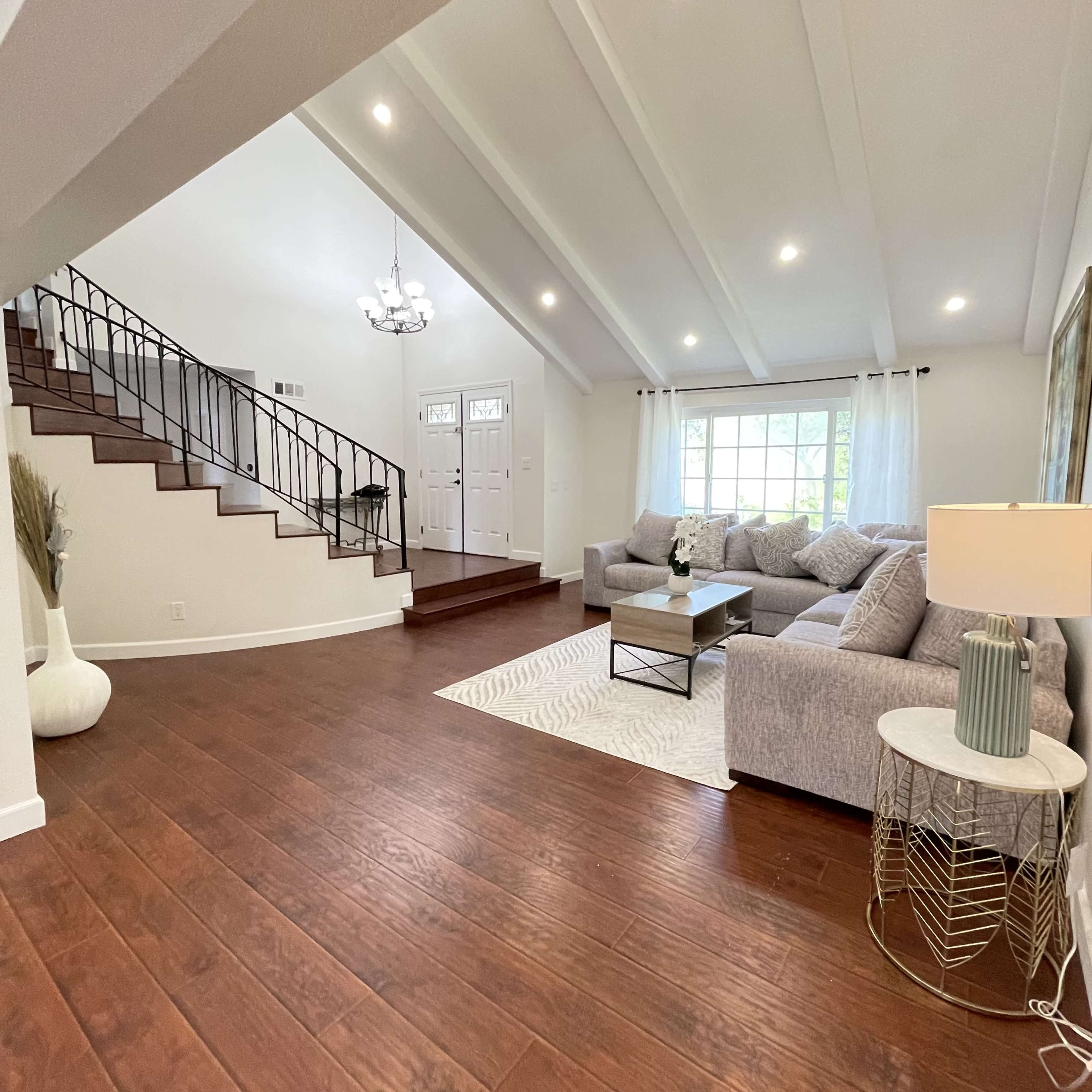 The image shows a spacious living room with a gray sectional sofa, a coffee table, and a patterned rug, featuring large windows and a staircase in the background.