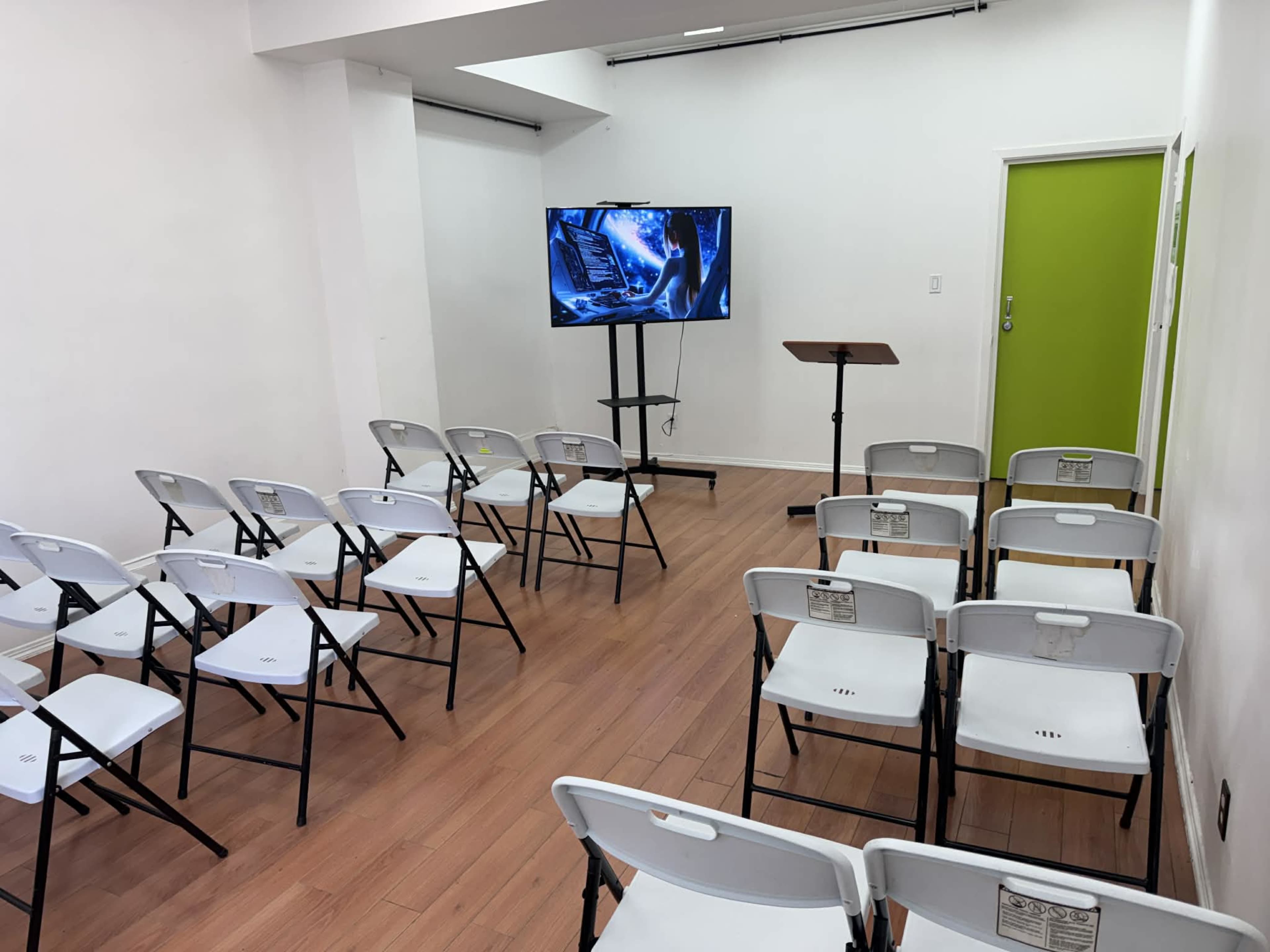 The image shows a small seminar room with chairs arranged in rows facing a TV on a stand and a podium against a white wall with a green door.