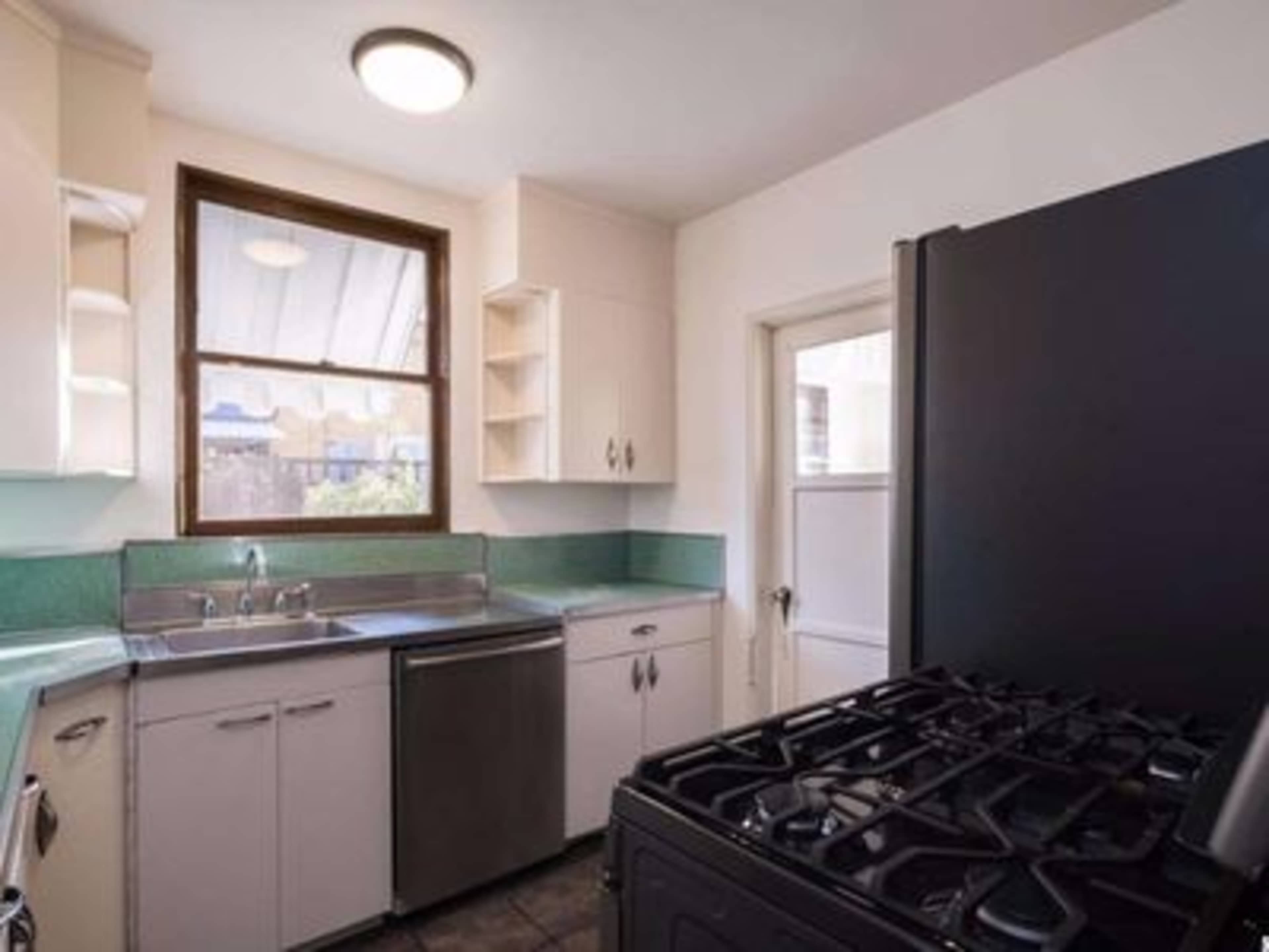 A small kitchen with a sink, a stove, and a door leading outside, illuminated by natural light from a window.