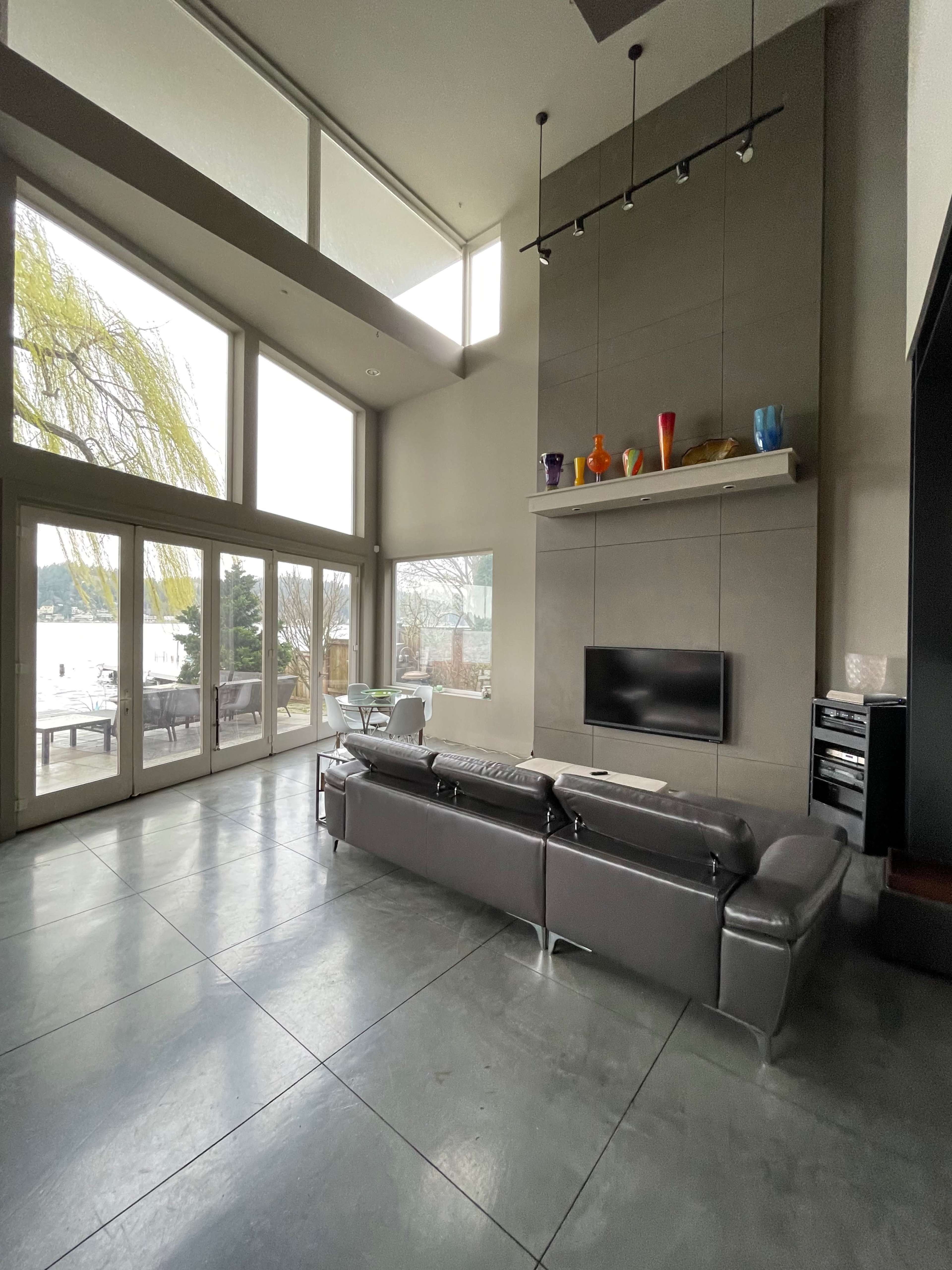 The spacious living room features large windows overlooking a lake, a modern silver couch, and a simple fireplace with colorful decor above it.