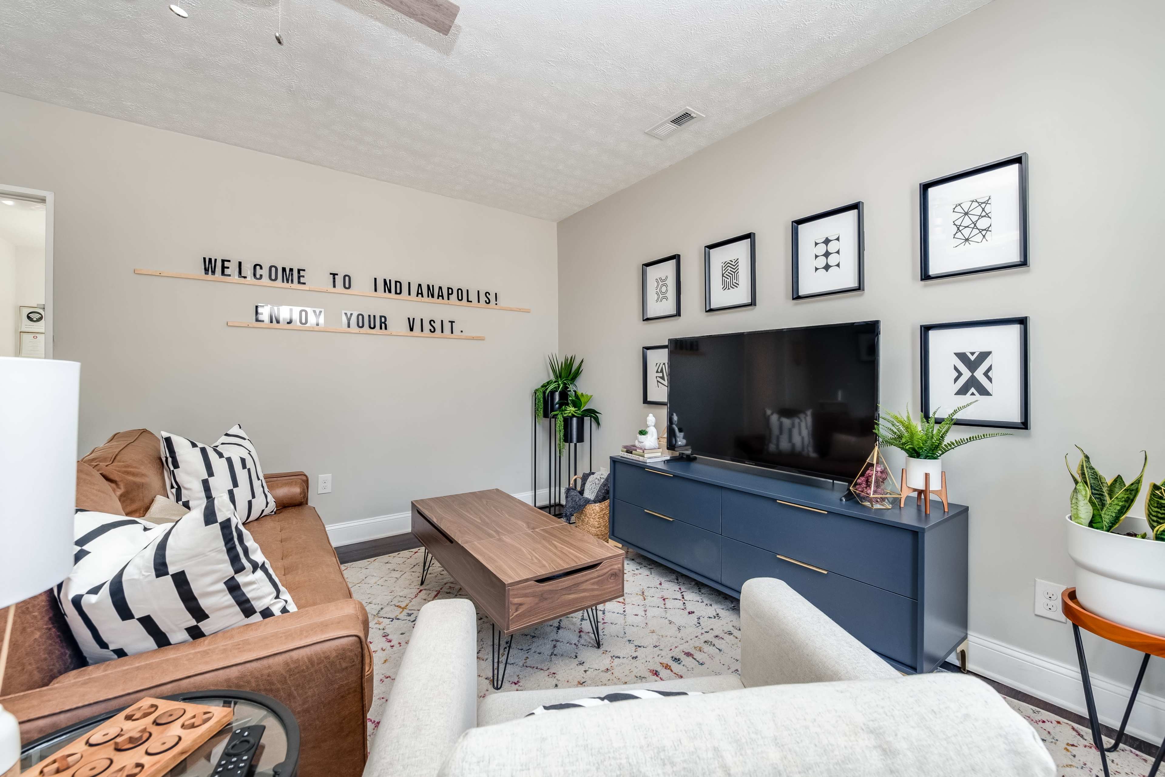 A cozy living room features a brown sofa, a gray TV stand, and framed artwork on the walls, with a sign that reads "WELCOME TO INDIANAPOLIS! ENJOY YOUR VISIT."