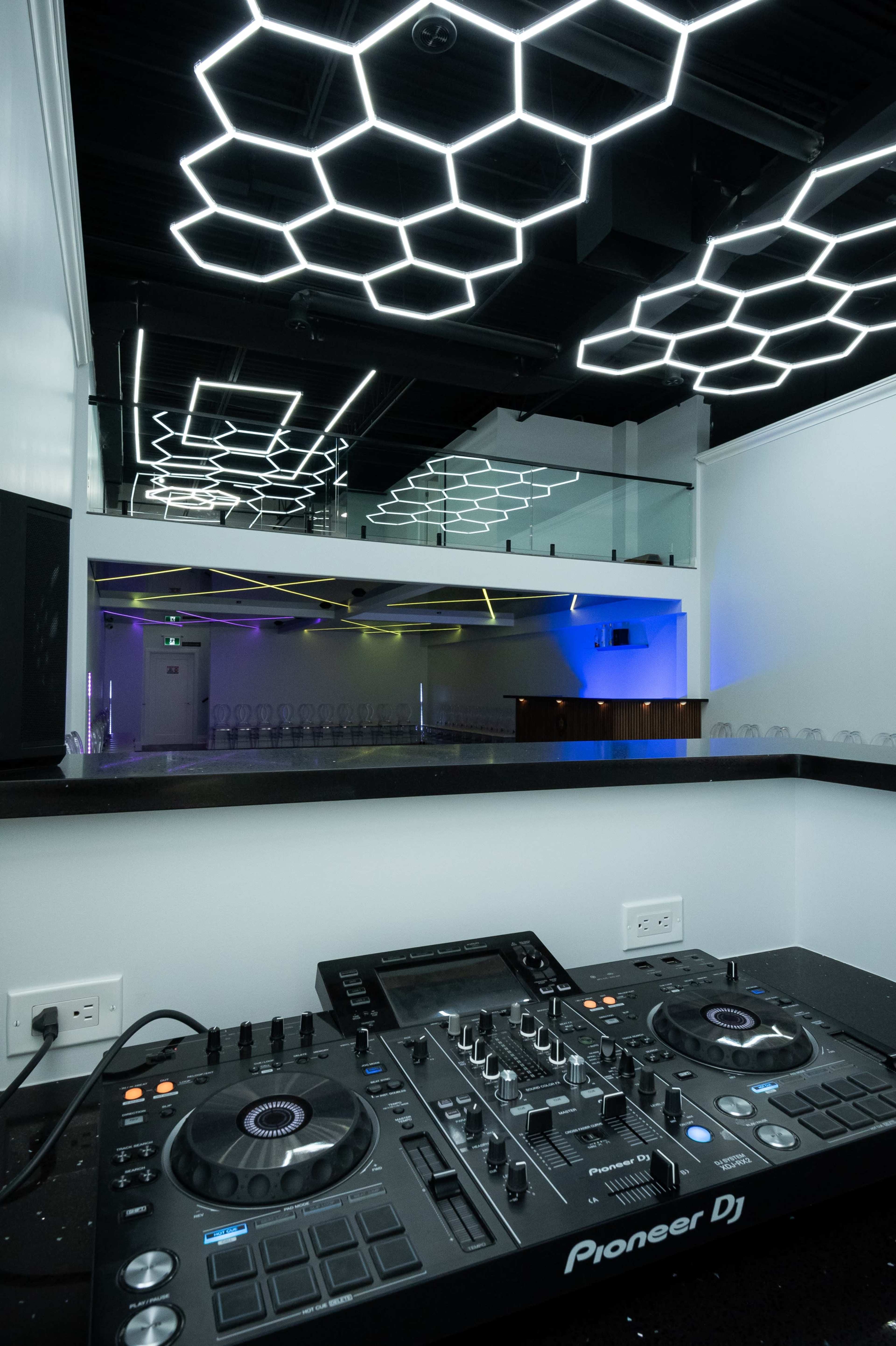 A modern DJ setup with a Pioneer DJ console in the foreground and a brightly lit, hexagon-patterned ceiling in an empty venue.