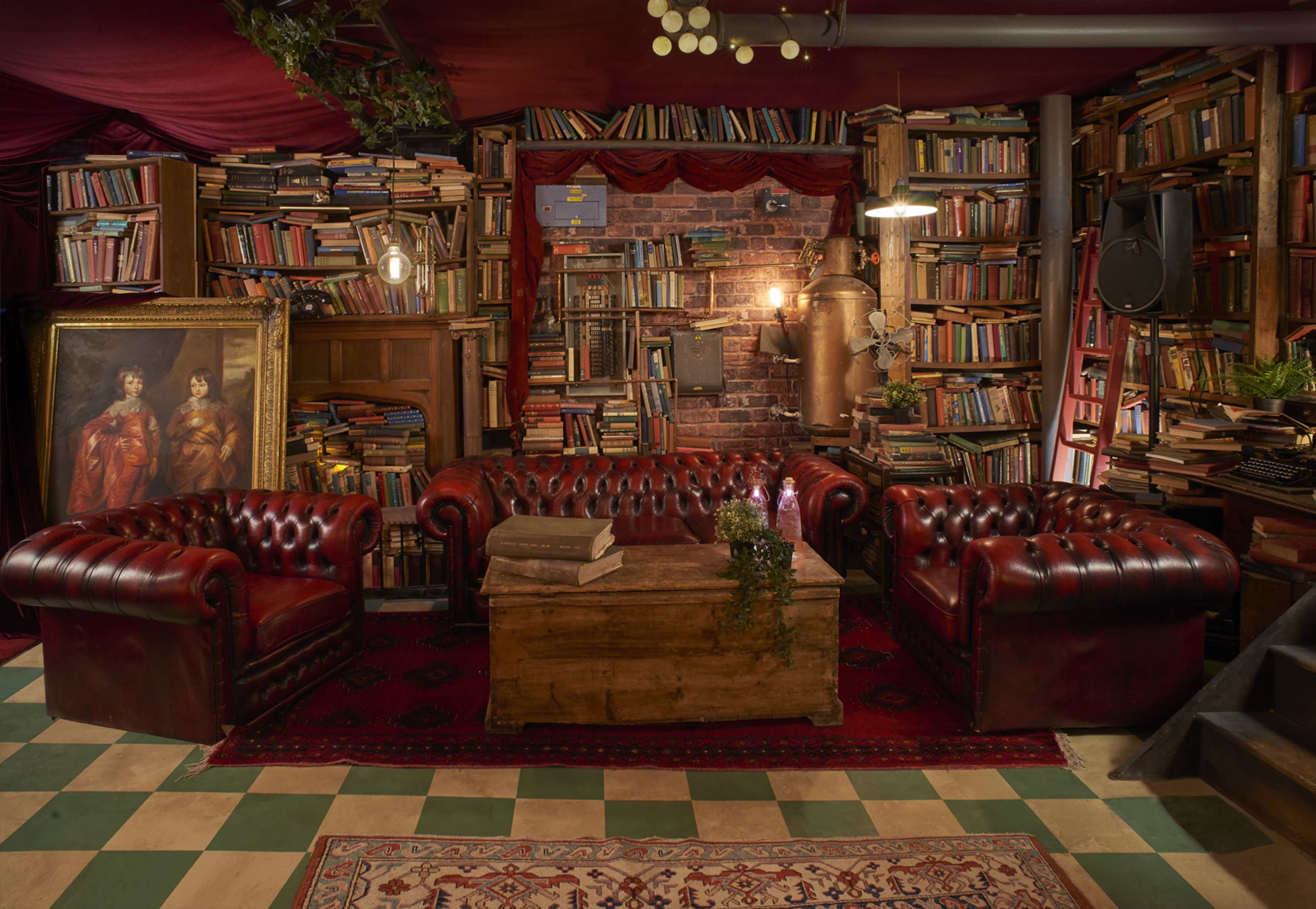 The image shows a cozy reading room featuring red leather chesterfield sofas, a wooden coffee table, and walls lined with bookshelves filled with books.