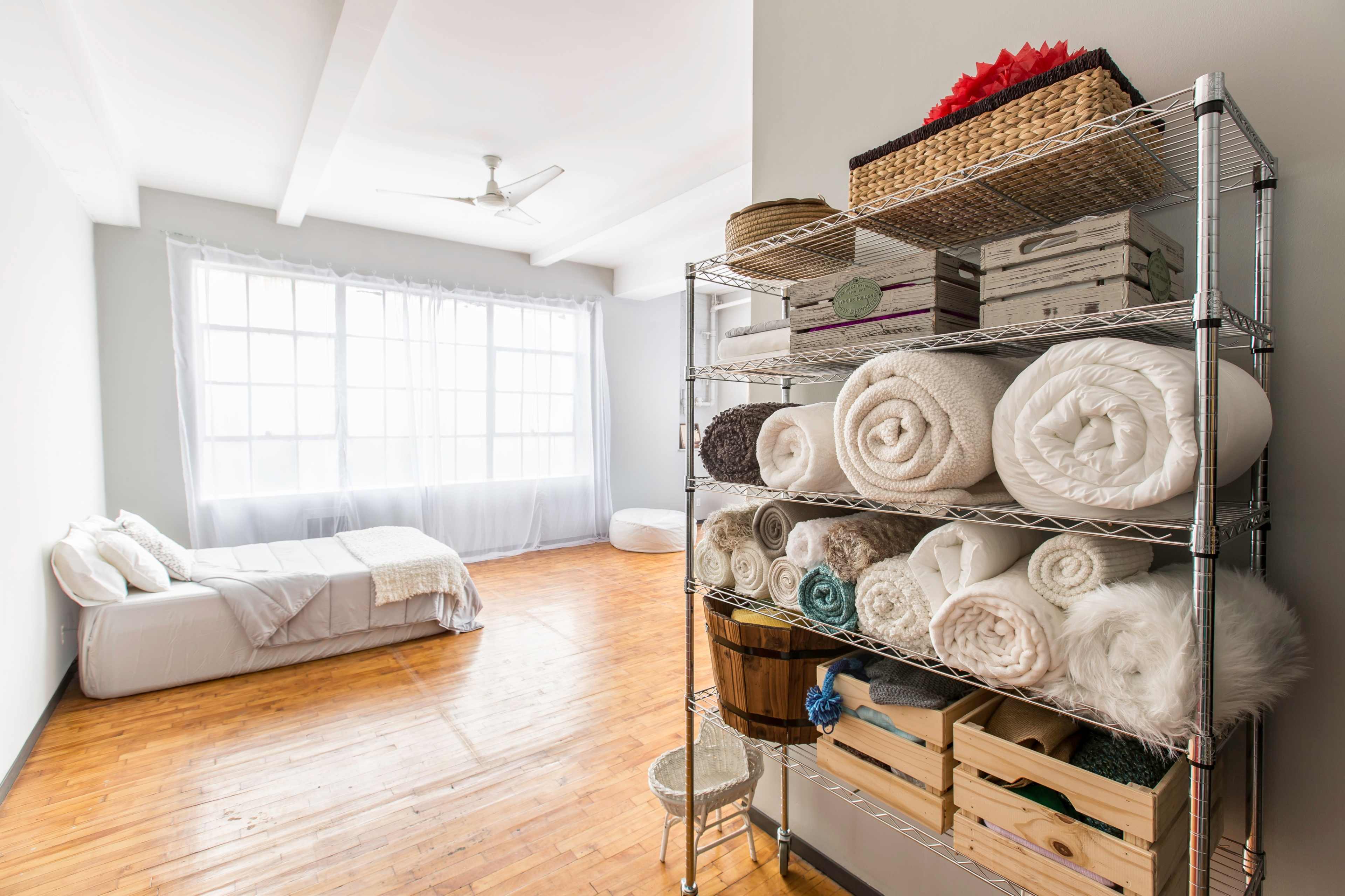 The image shows a minimalist bedroom featuring a neatly organized shelf of folded blankets and towels, with a bed and large windows in the background.