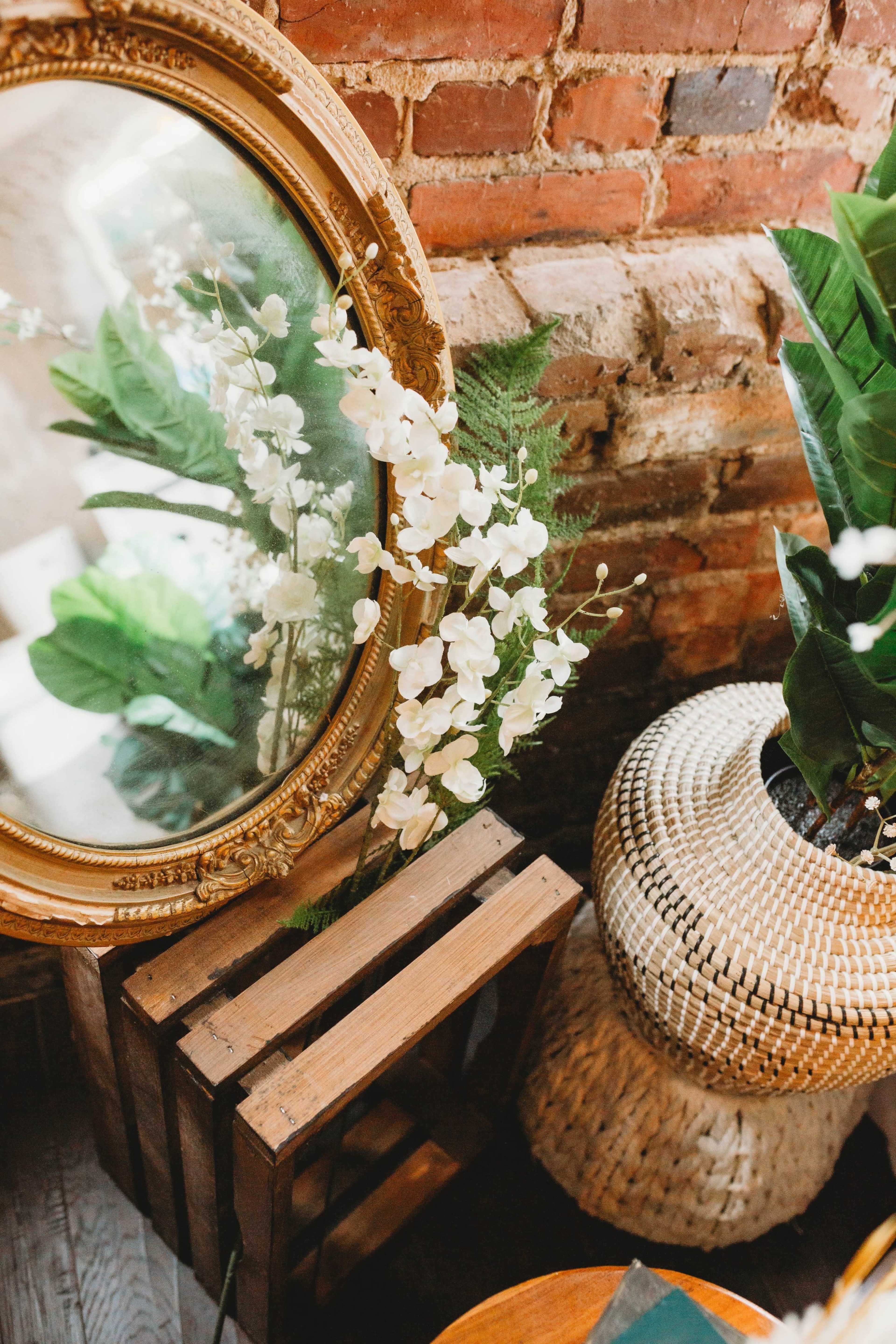 A large, ornate mirror reflects a bouquet of white flowers beside a woven basket plant holder in a room with exposed brick walls.