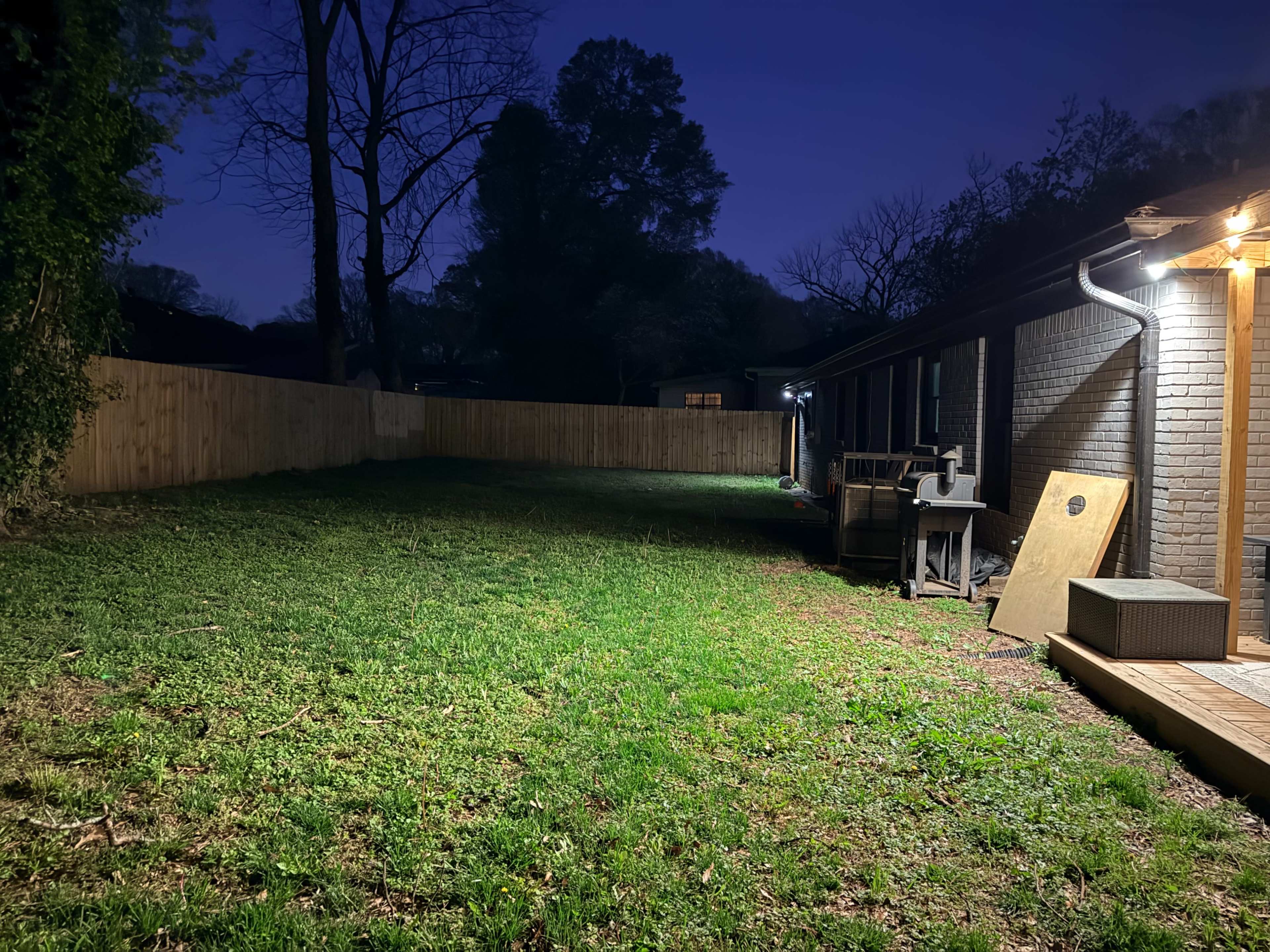 The image shows a dimly lit backyard with grass, a wooden fence, and several objects near the house.