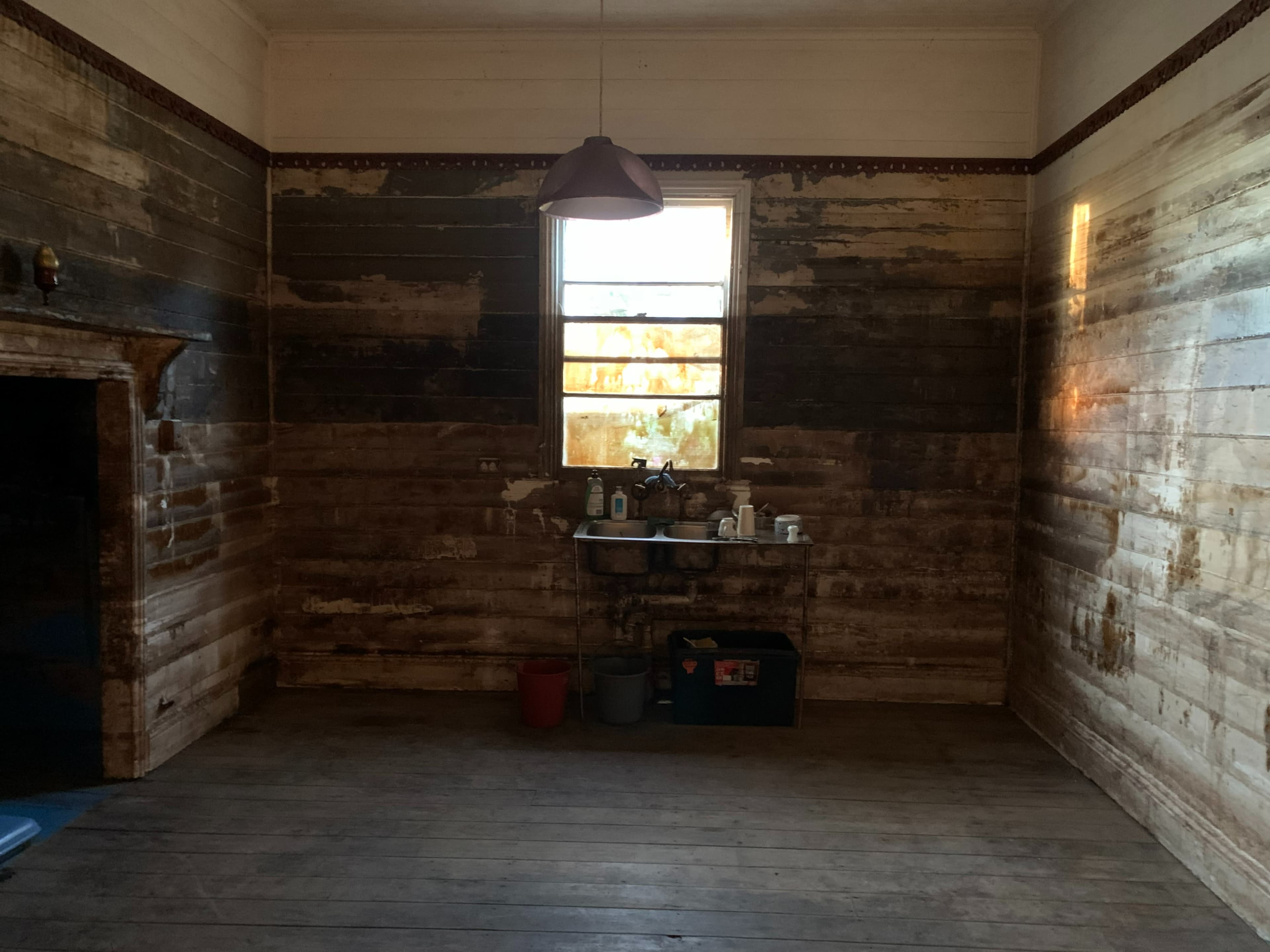 The image shows an empty room with weathered wooden walls, a window letting in light, and a small kitchen area with basic utensils and containers.