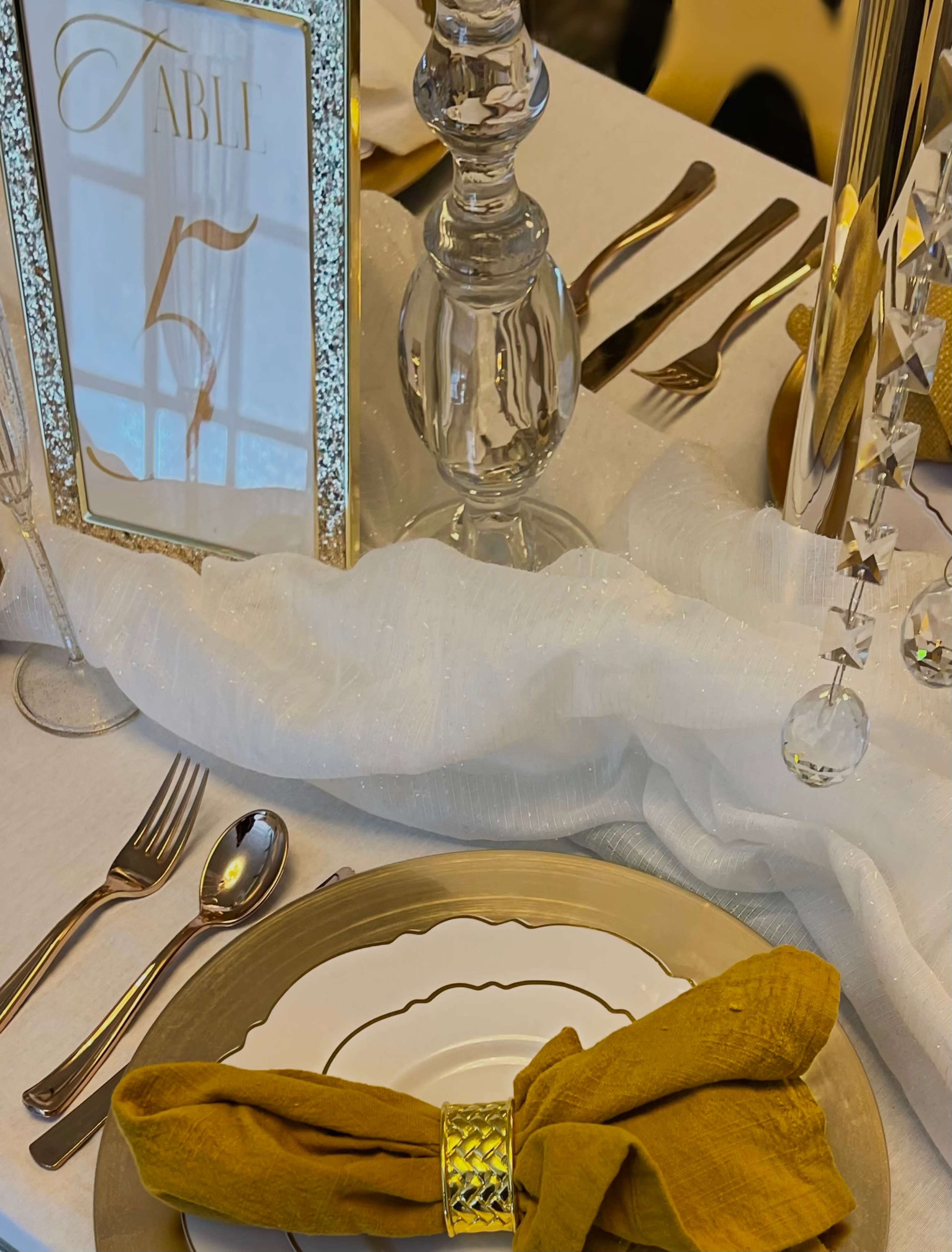 A elegantly set dining table featuring a gold and white color scheme, with a centerpiece displaying a framed sign for "Table 5," along with polished cutlery and decorative napkins.