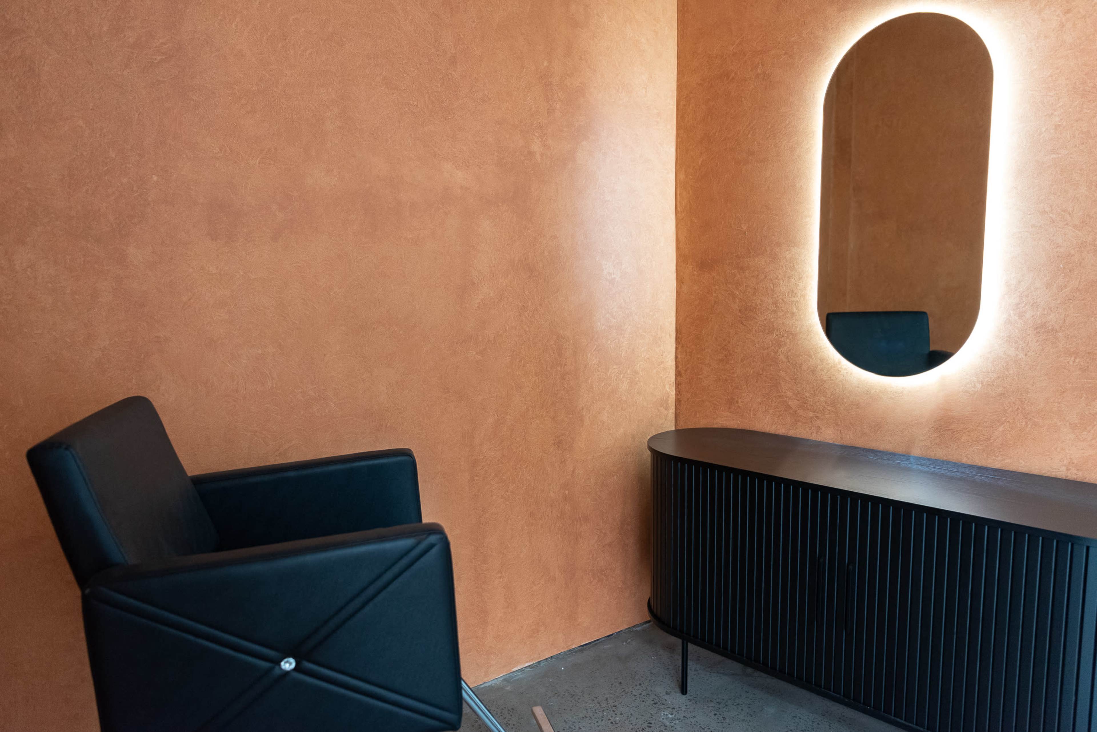 A minimalistic hair salon interior with an orange textured wall, a black chair, a sleek black vanity, and a rounded mirror with an integrated light.
