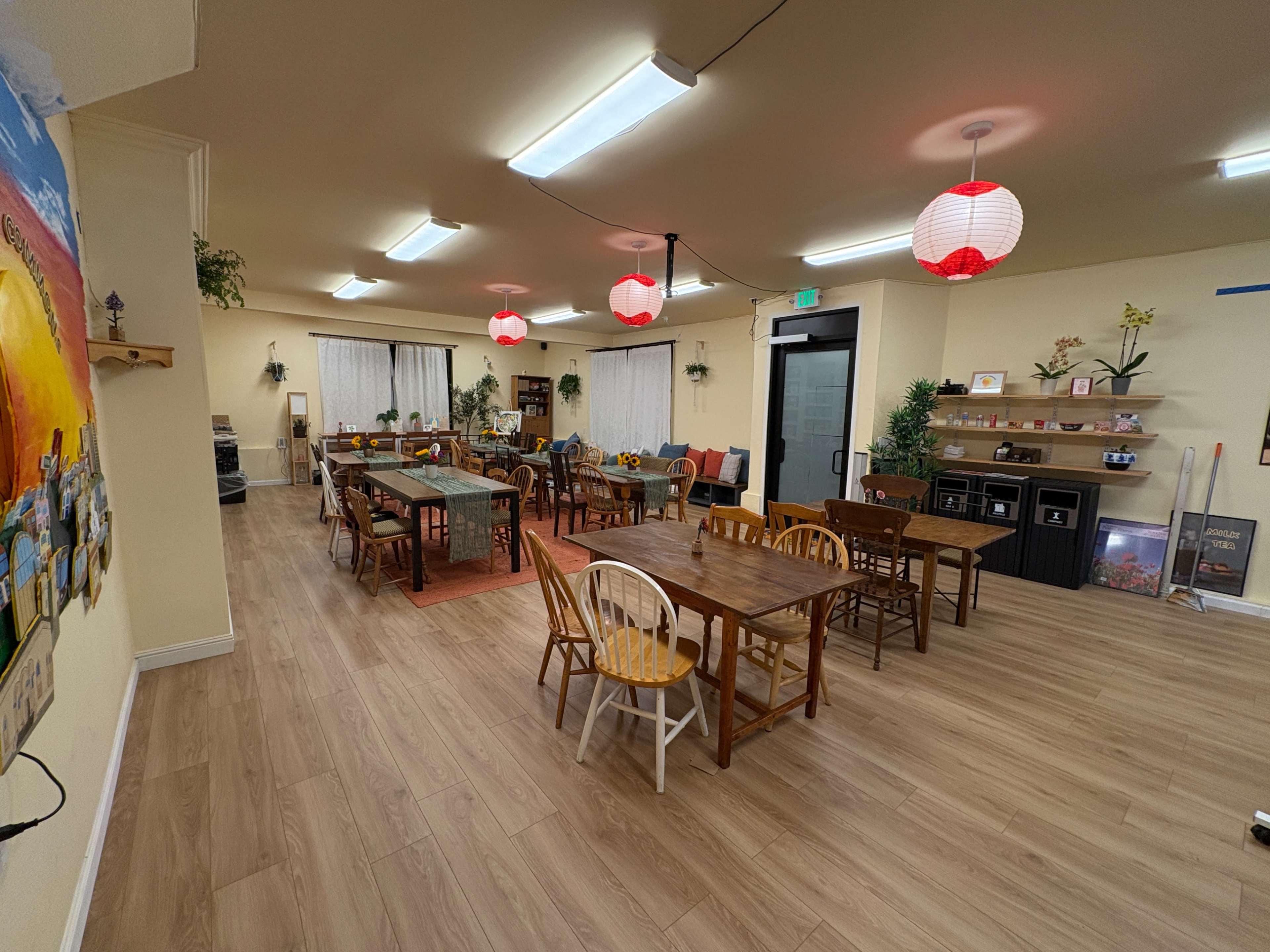 The image shows a spacious indoor area with multiple wooden tables and chairs, bright lighting, and decorative elements like lanterns and plants.