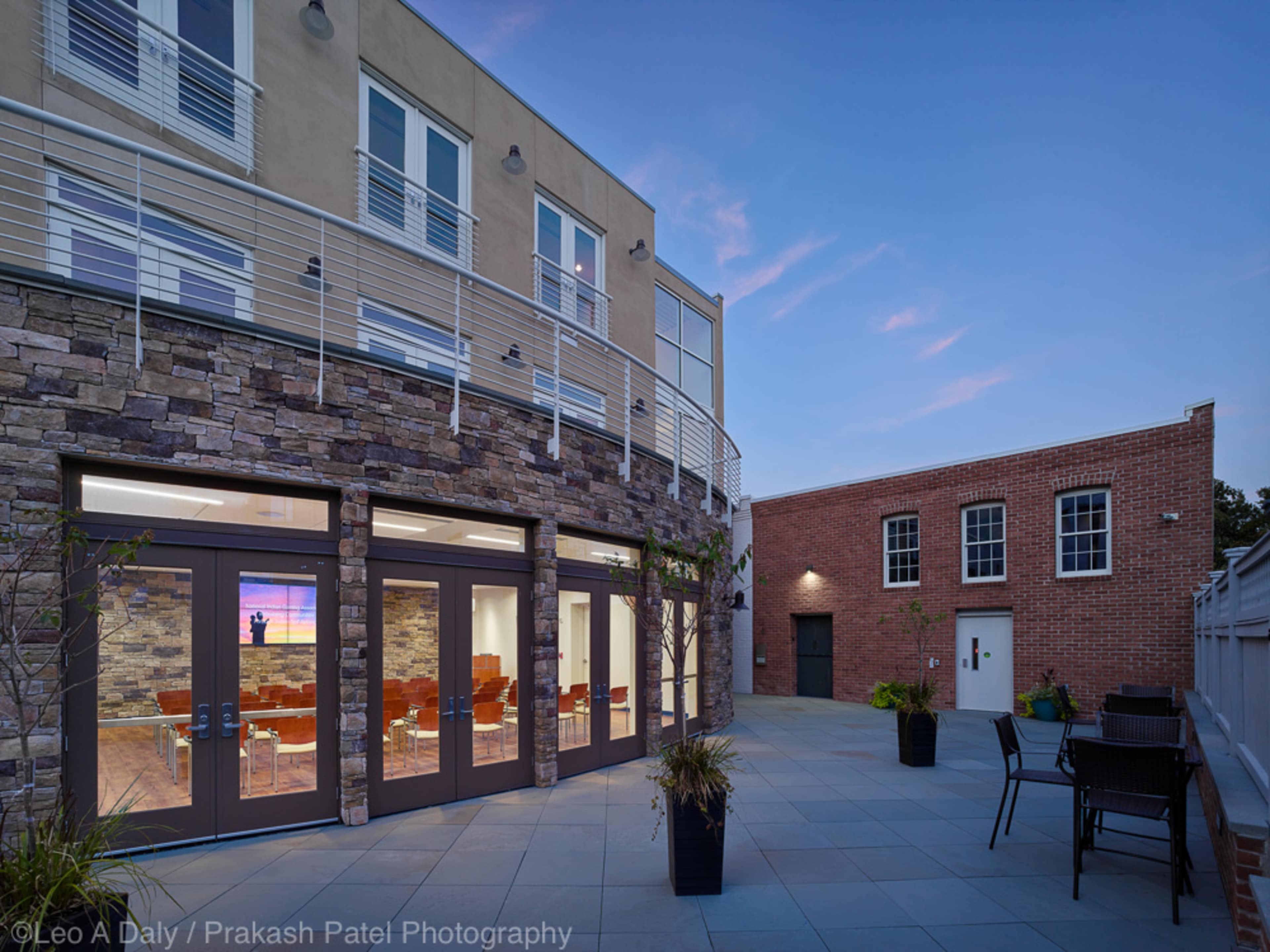 A modern building with stone walls and large glass doors leading to a patio area featuring outdoor furniture against a twilight sky.