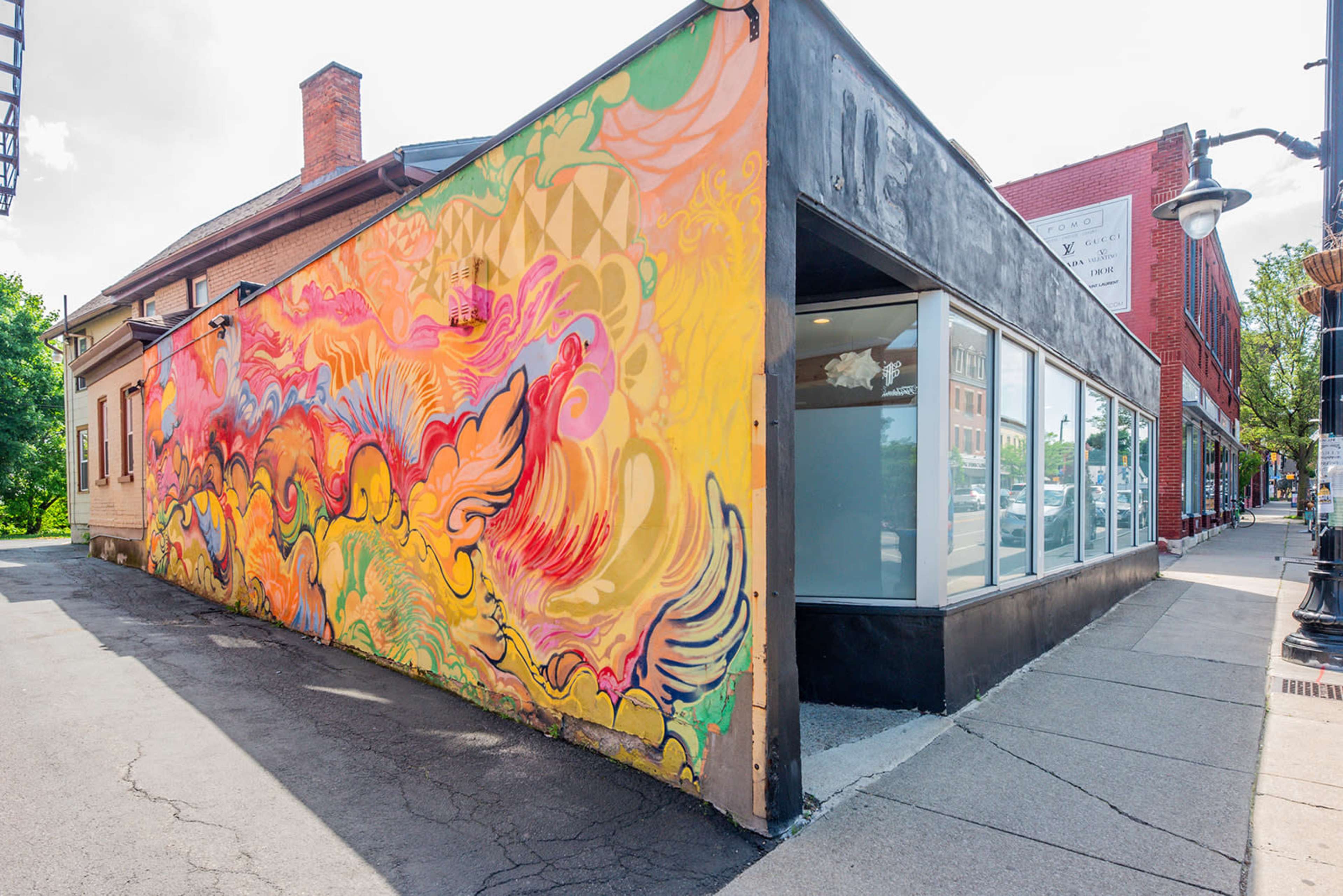 A colorful mural adorns the side of a building, extending around the corner onto a sidewalk beside a street.