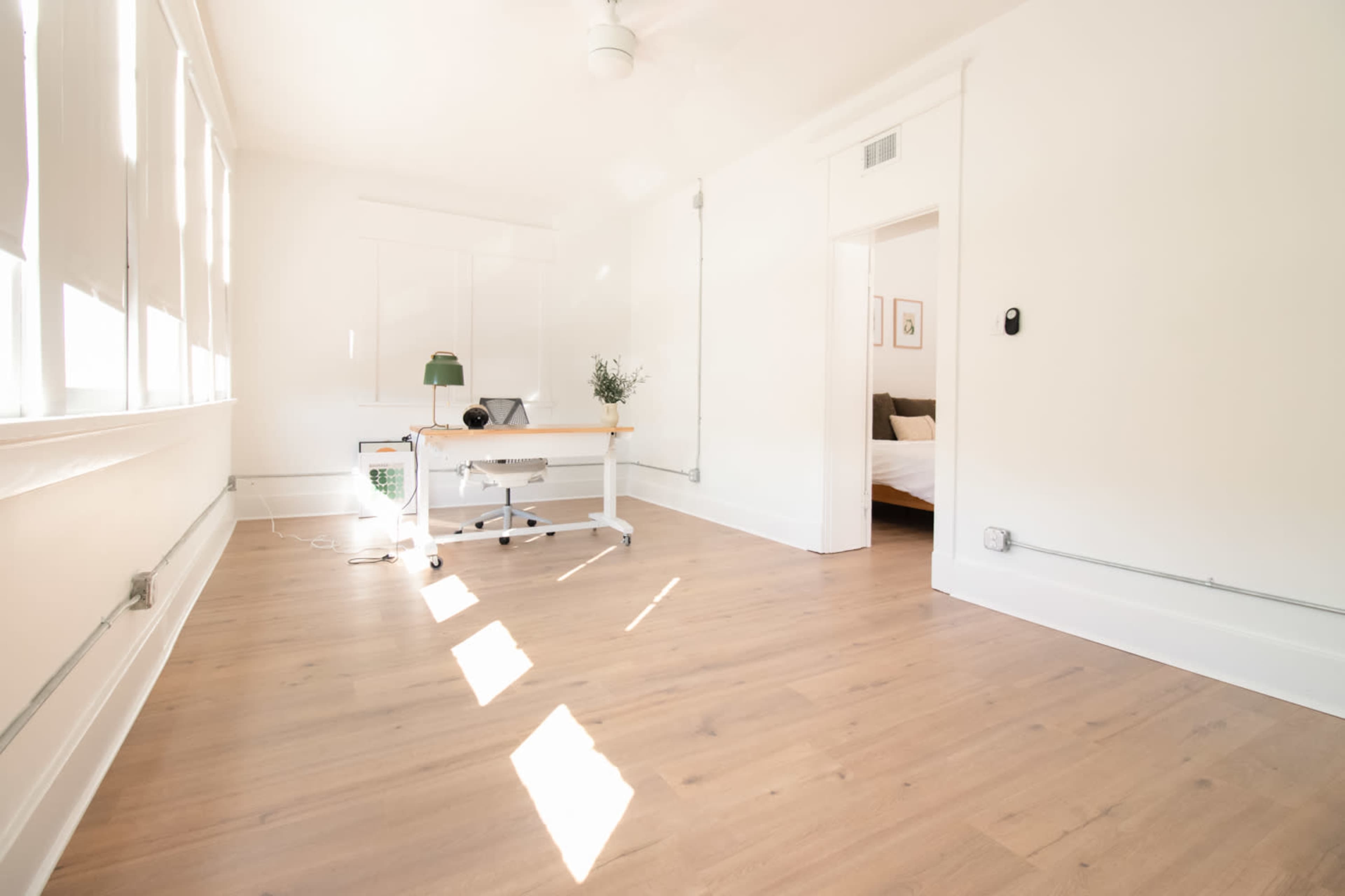 A bright, minimalist room features a wooden desk and a lamp, with sunlight streaming through several windows.
