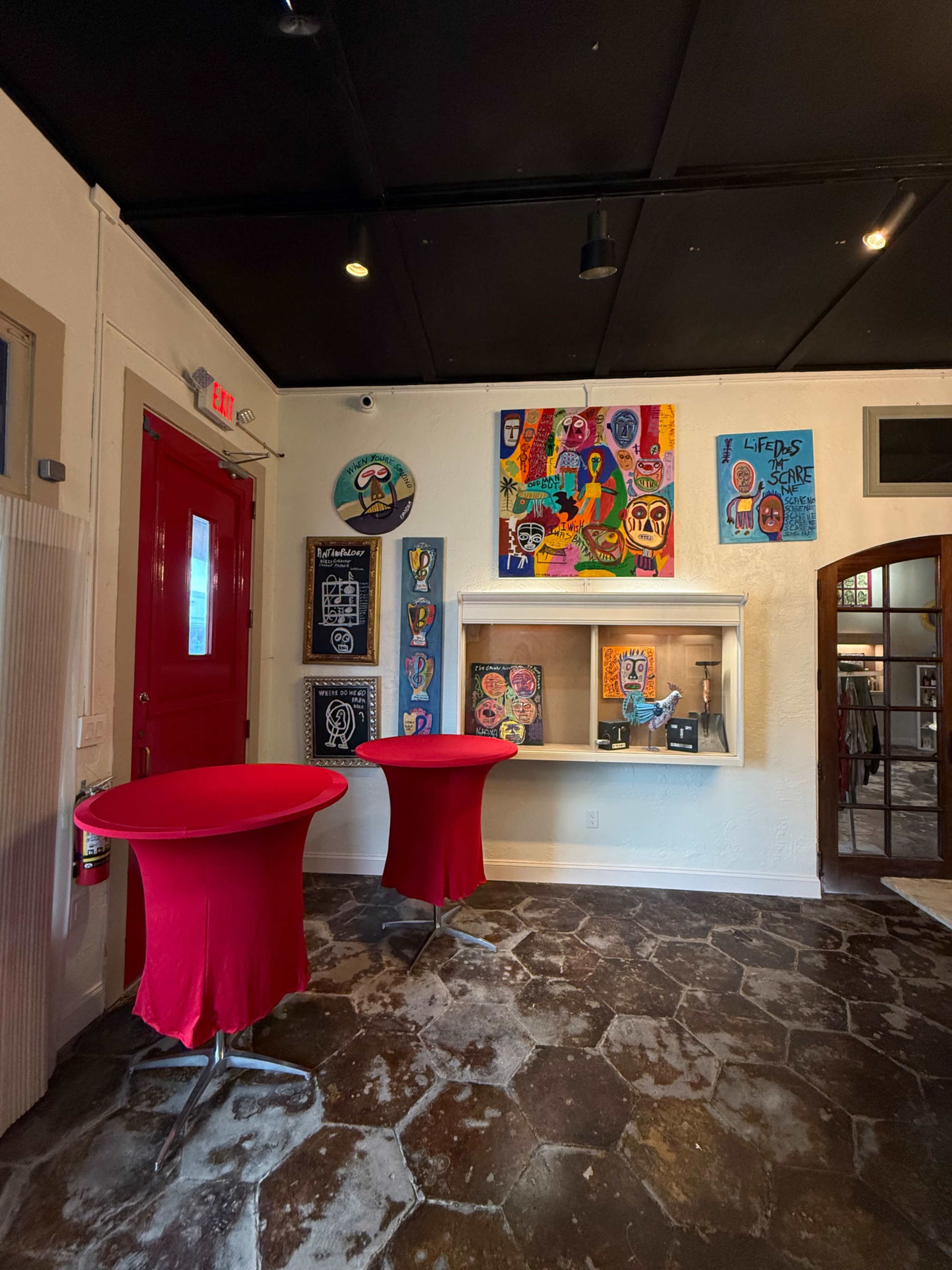 The scene features a brightly colored room with two red cocktail tables and walls adorned with various pieces of art, including colorful paintings, against a backdrop of stone flooring.
