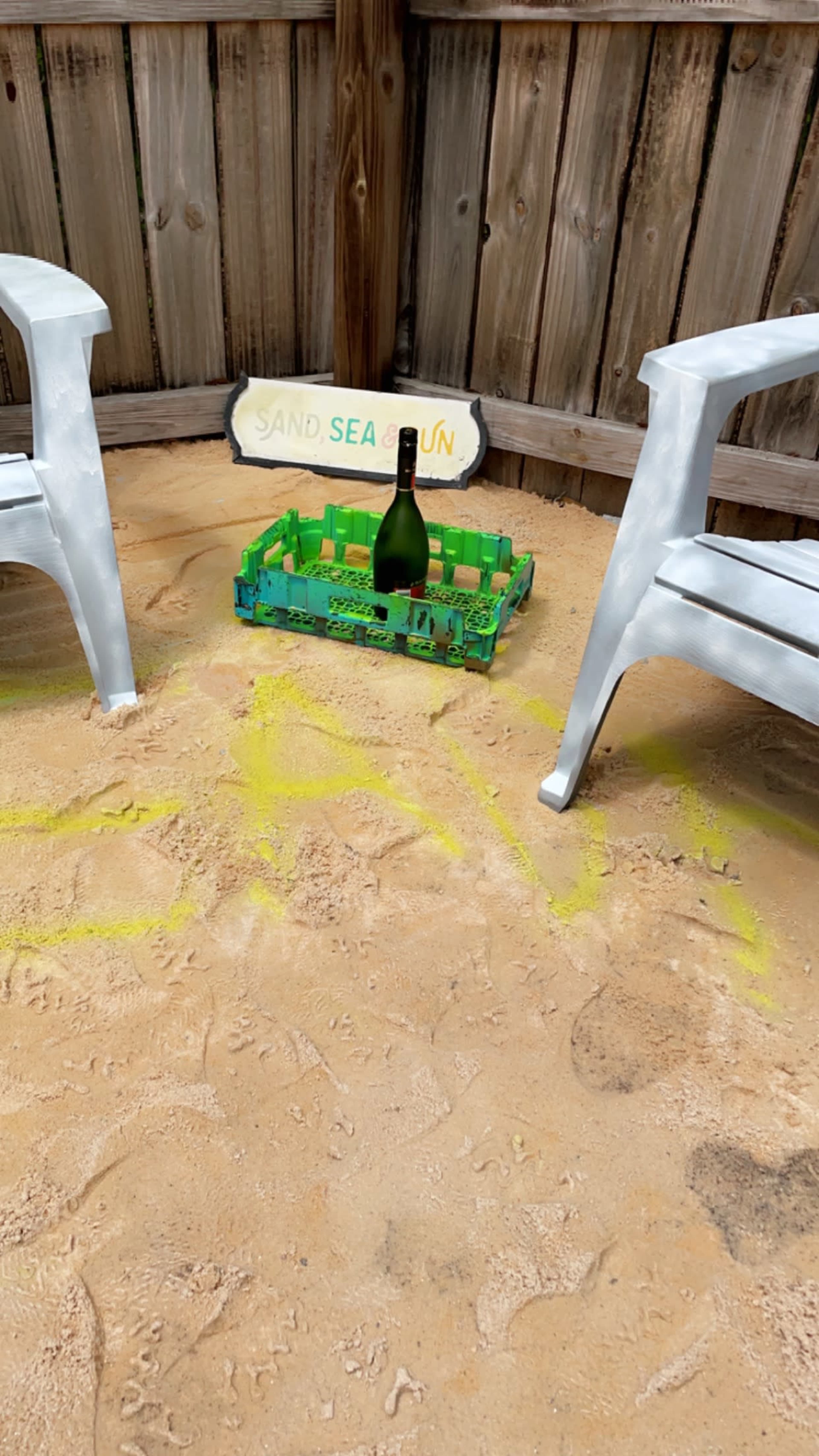 A sandy area enclosed by wooden fencing, featuring two white chairs and a green tray holding a bottle of champagne.