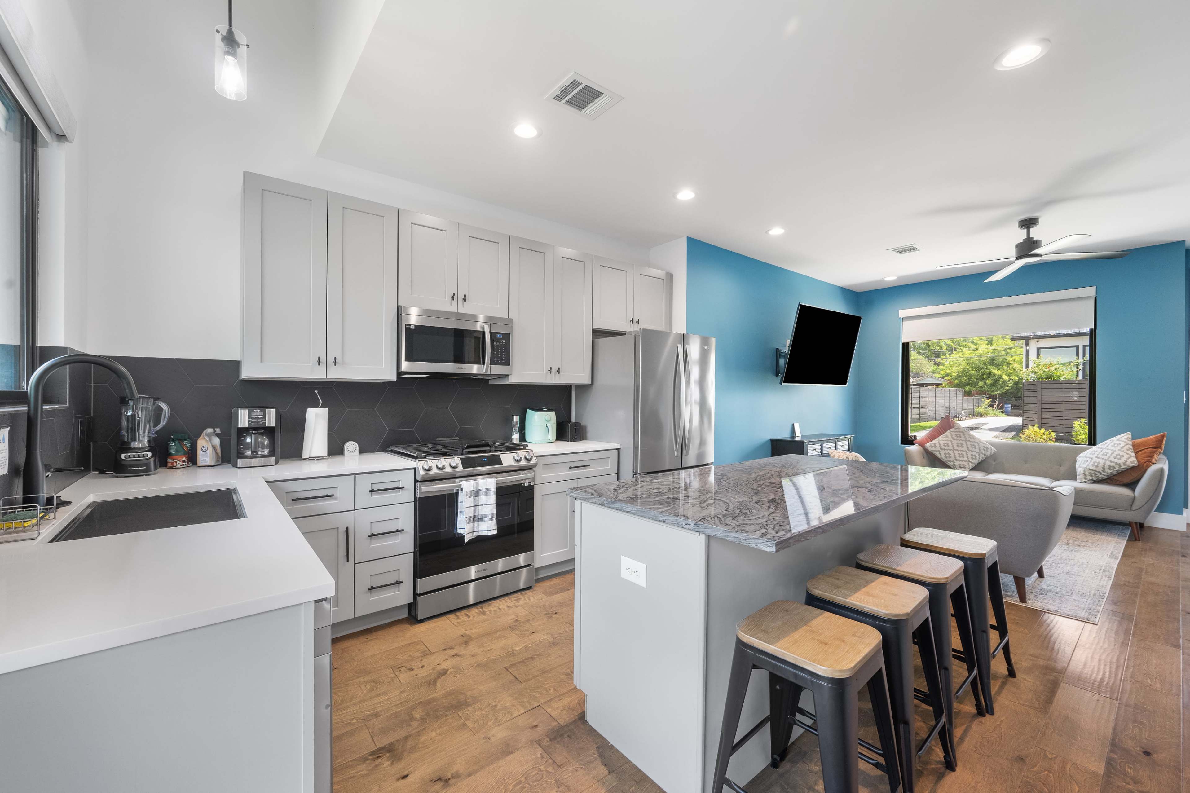 The image shows a modern kitchen and living area with gray cabinetry, stainless steel appliances, a central island with bar stools, and a blue accent wall.