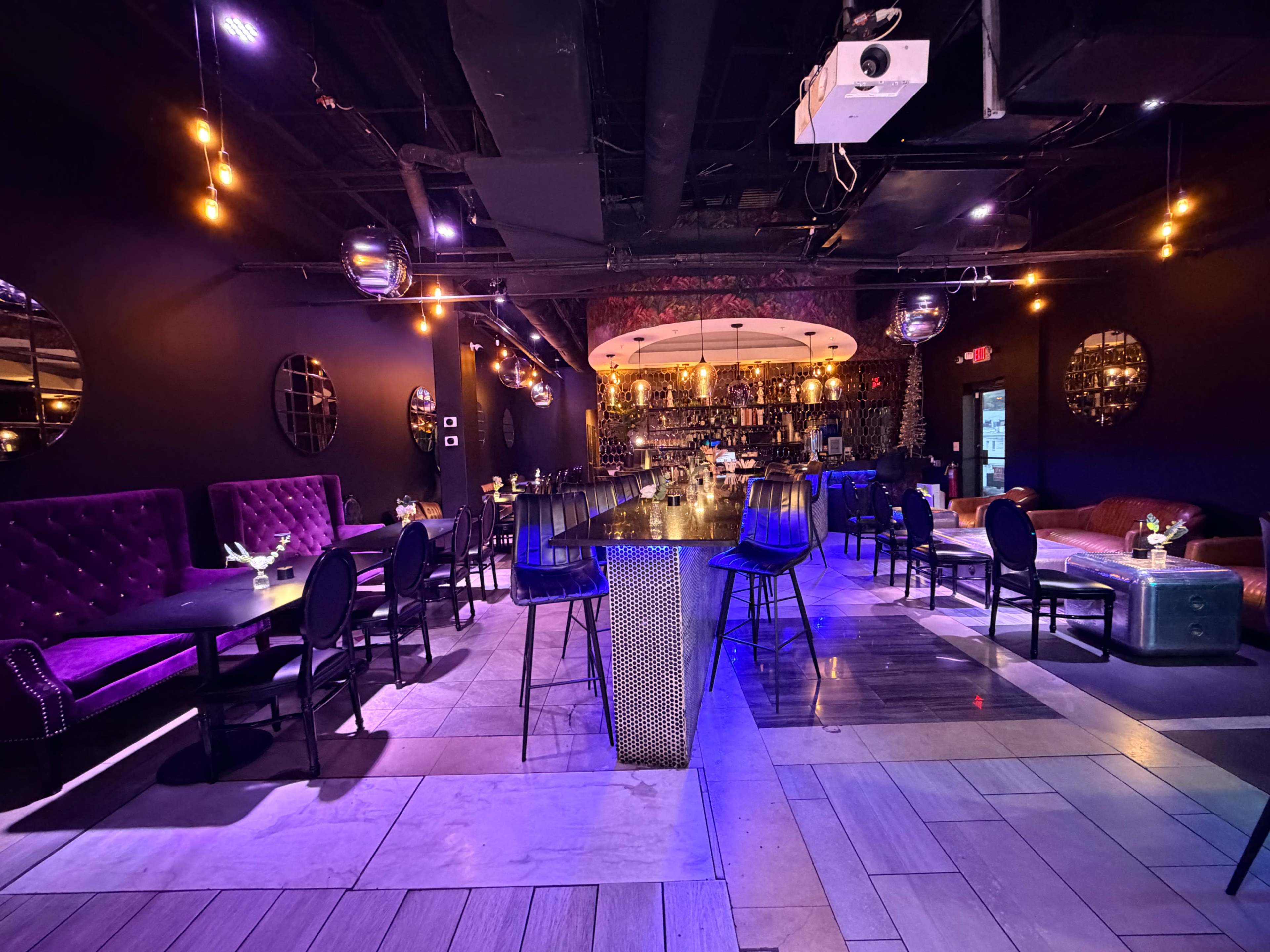 The image shows a dimly lit bar and dining area with modern furnishings, featuring purple velvet seating and a long bar illuminated by pendant lights.