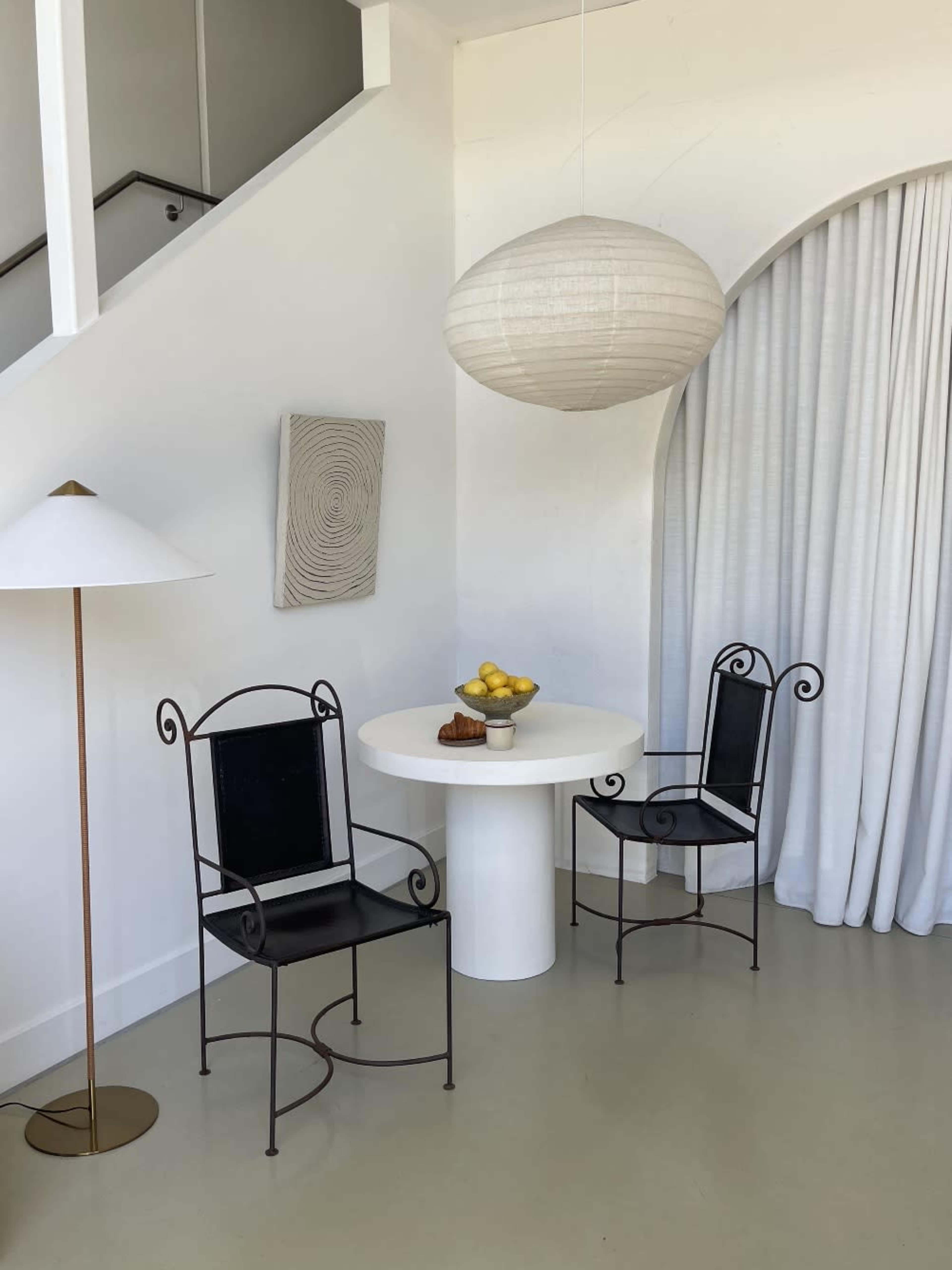 The image shows a minimalist interior featuring a round white table with two wrought iron chairs, a bowl of lemons, and a modern pendant light, all set against a white backdrop with curtains.