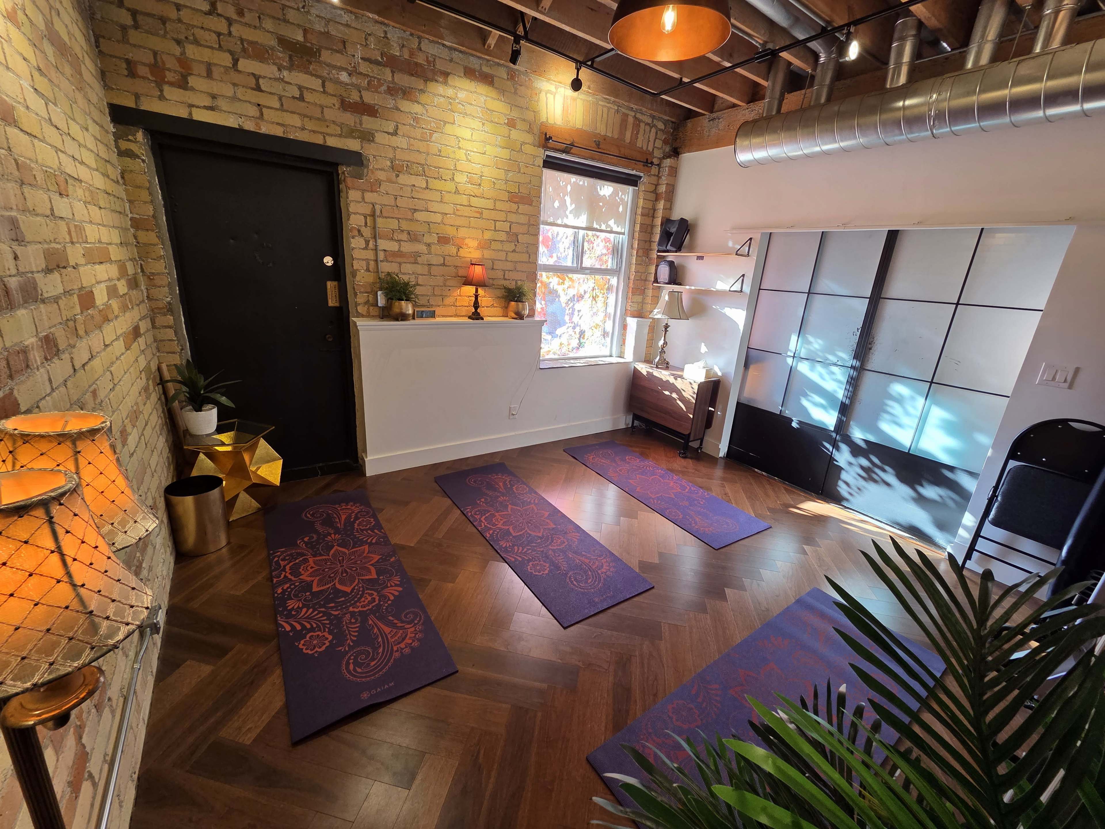 The image shows a yoga studio with three purple mats laid out on a wooden floor, surrounded by brick walls, plants, and warm lighting.