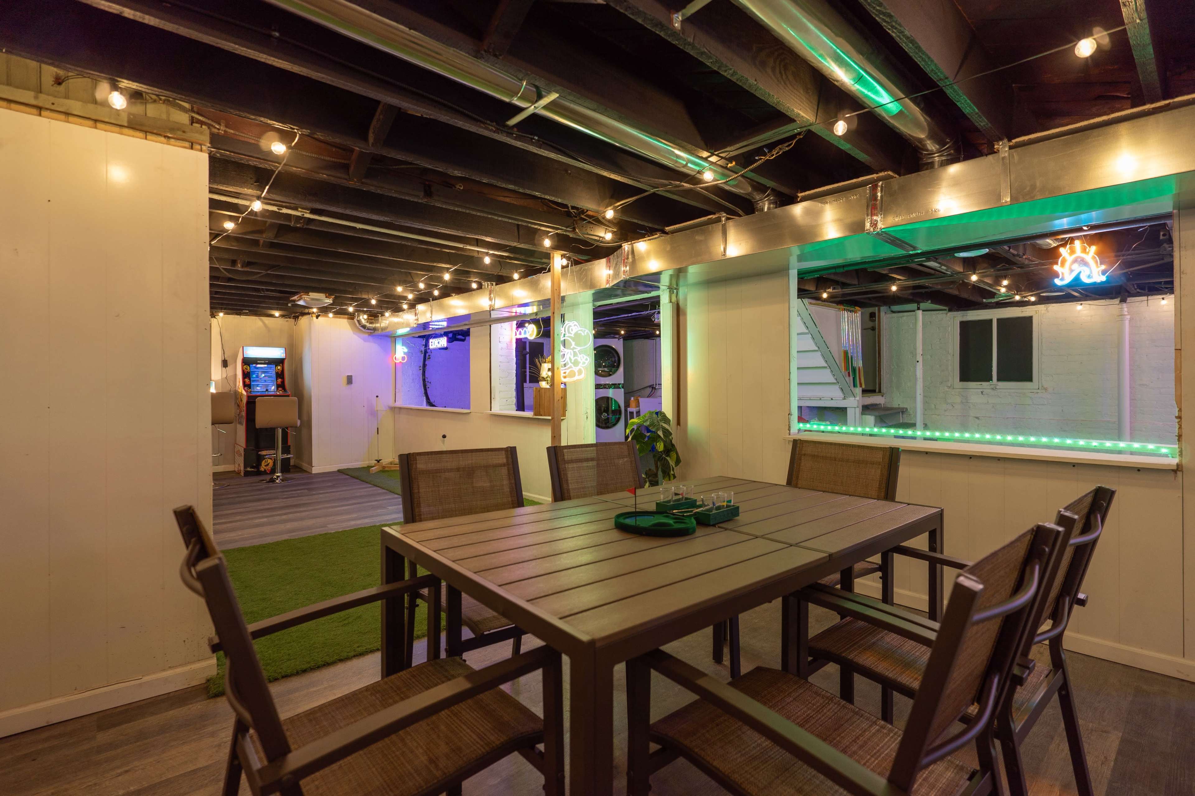 The image shows a spacious basement area with a dining table and chairs, illuminated by string lights and featuring neon signs in the background.