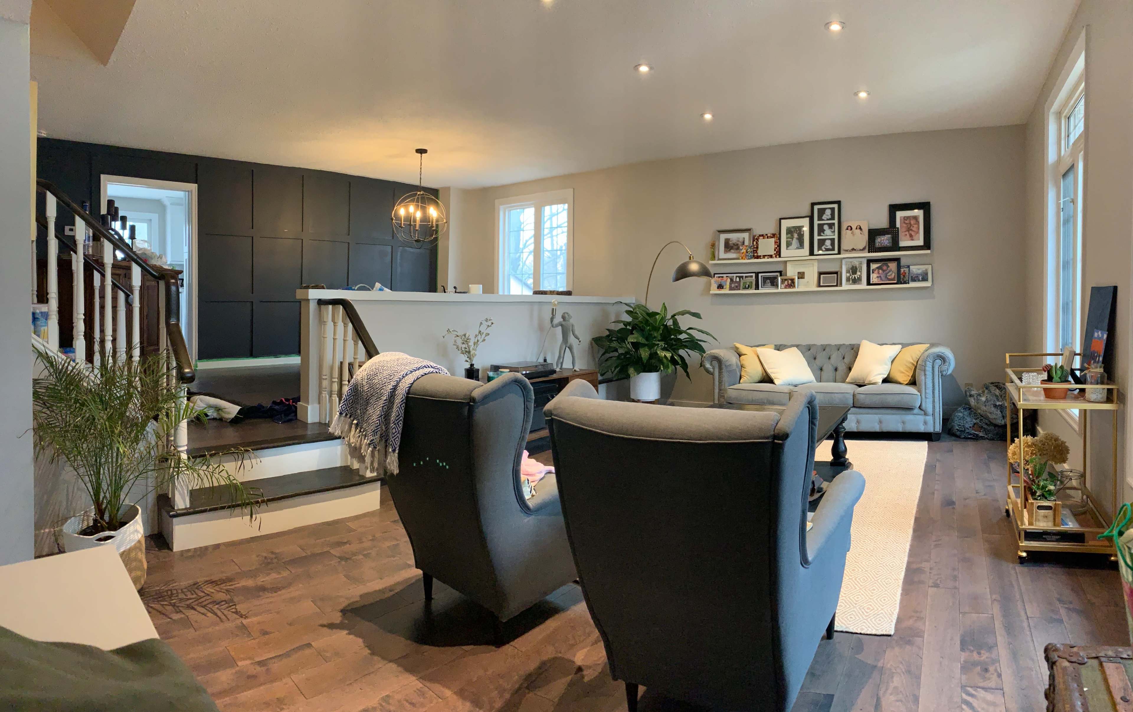 The image shows a modern living room with gray armchairs, a light-colored rug, and a staircase leading to a kitchen area in a spacious layout.