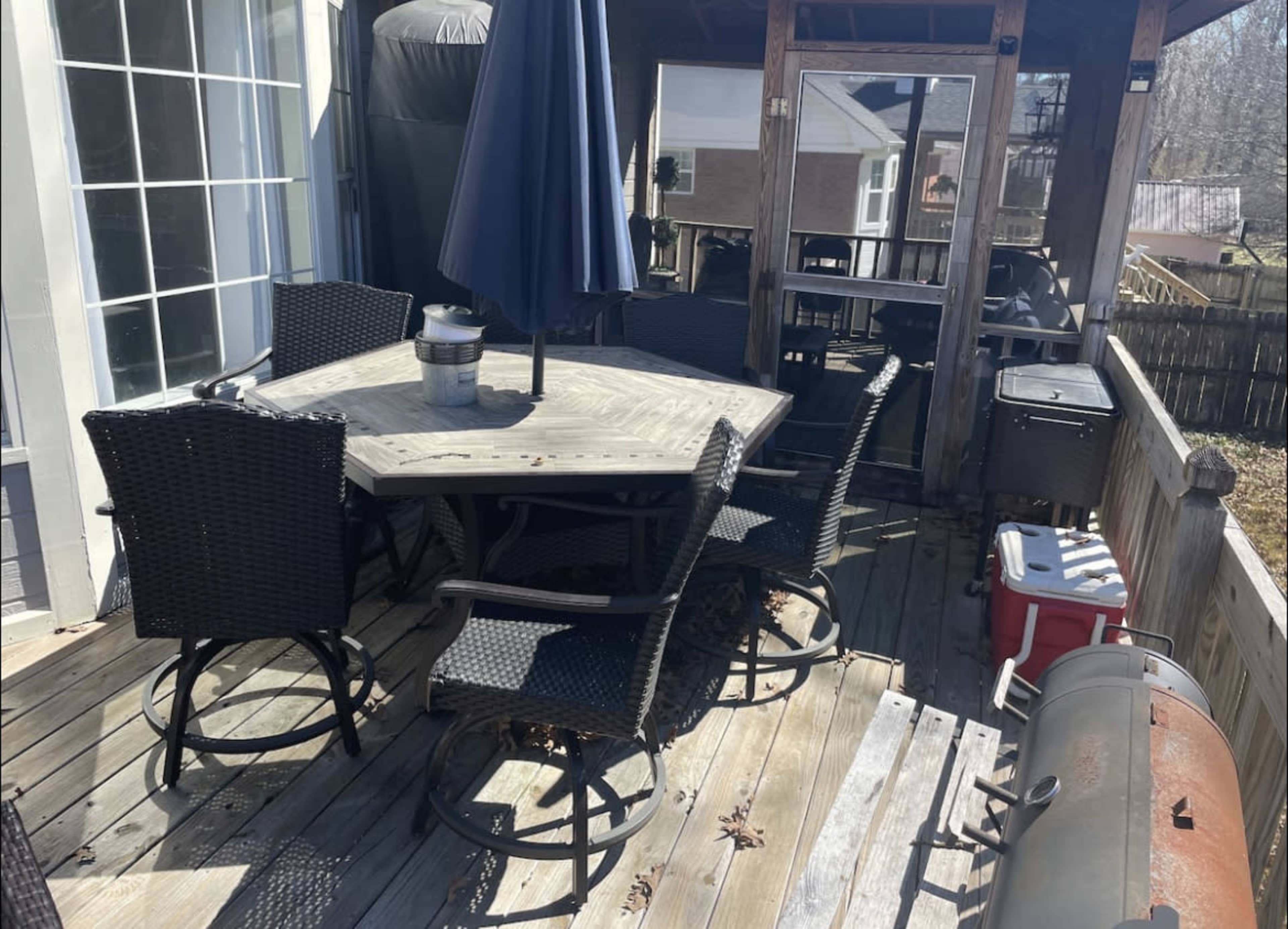 A wooden deck features an octagonal table surrounded by six wicker chairs, accompanied by a grill and a cooler in the corner.