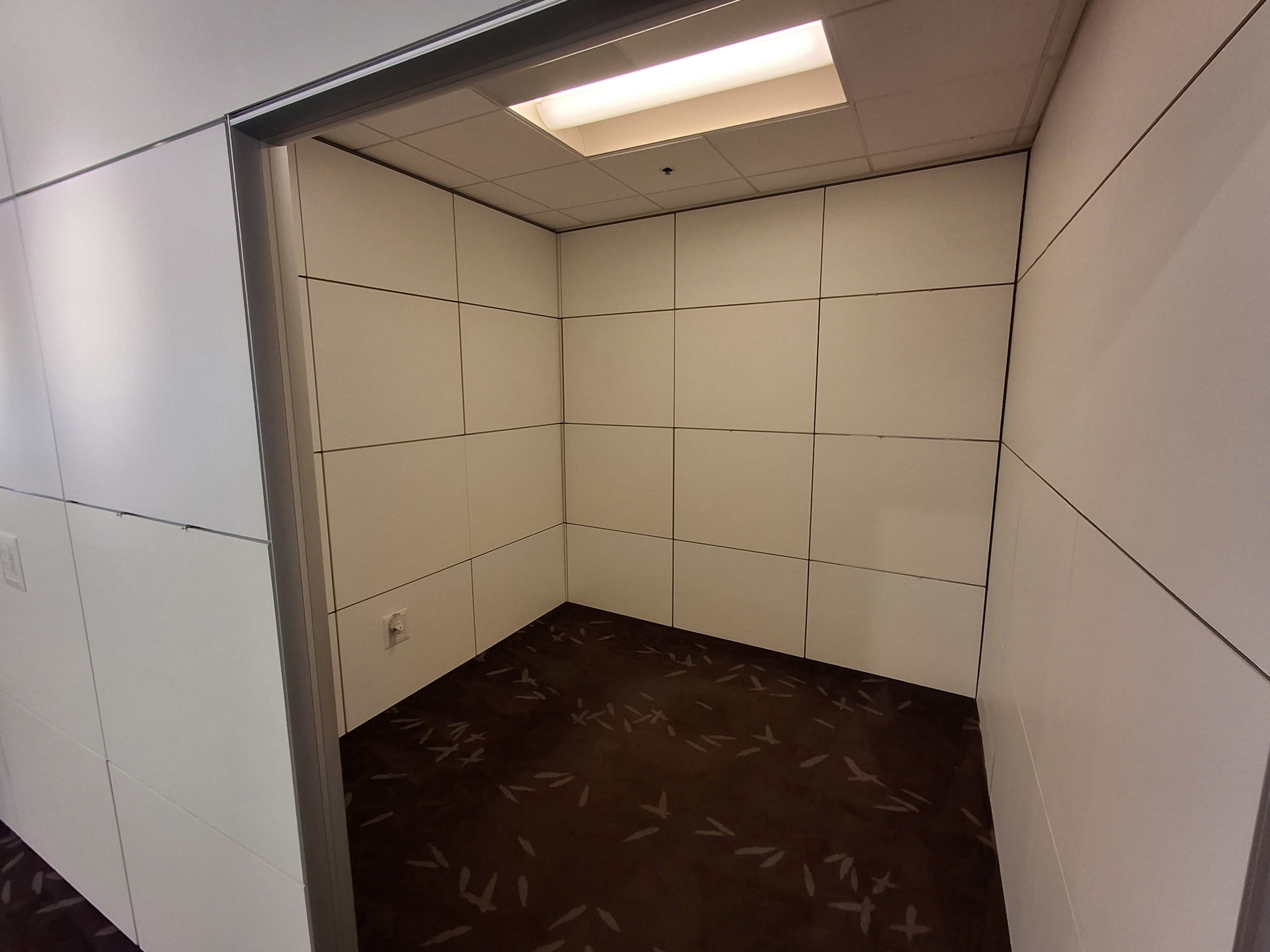 The image shows an empty, small room with plain white walls and a dark carpeted floor.
