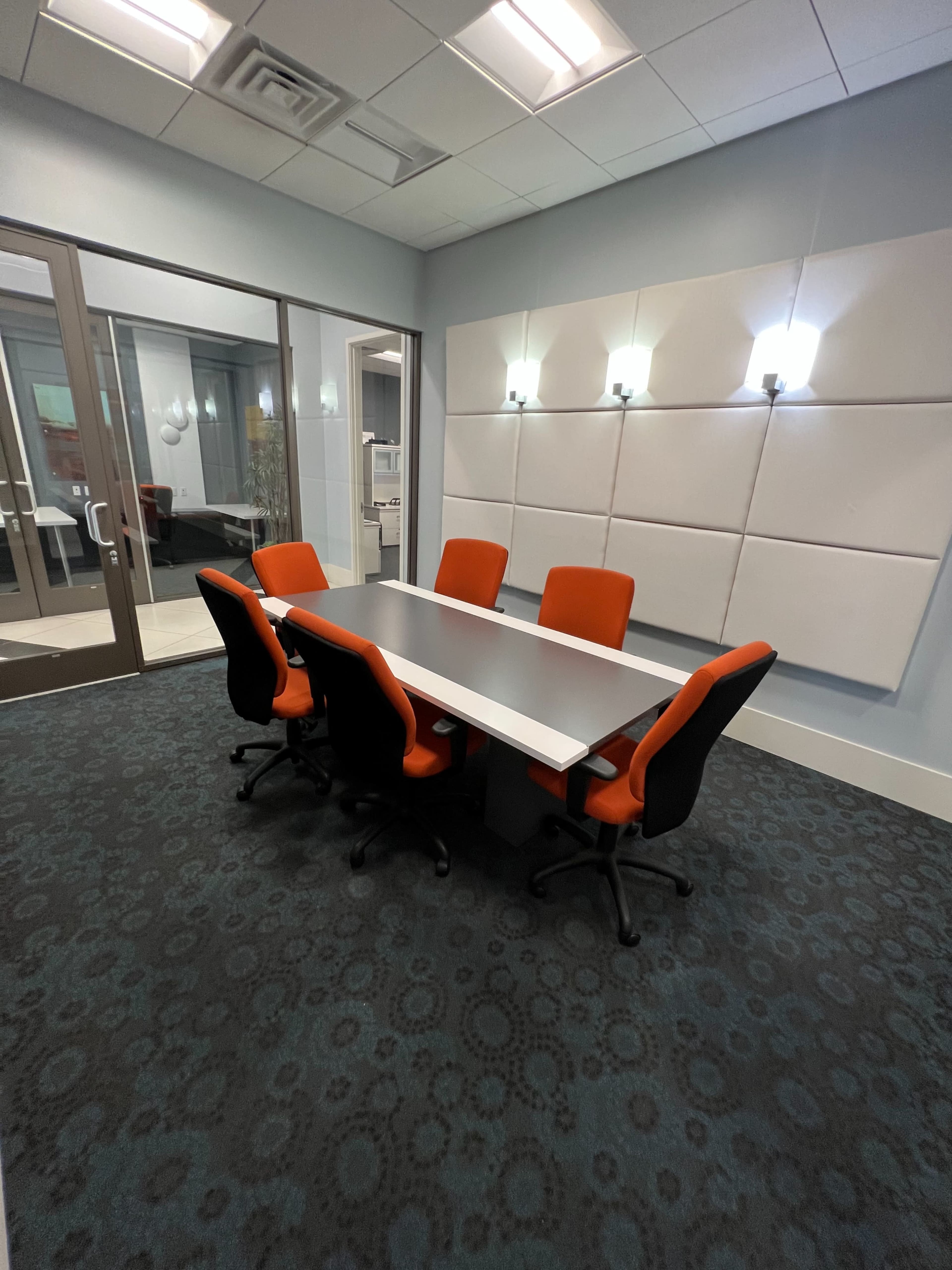 A modern conference room features a large gray table surrounded by black and orange chairs, with wall-mounted lights illuminating the white panel behind.