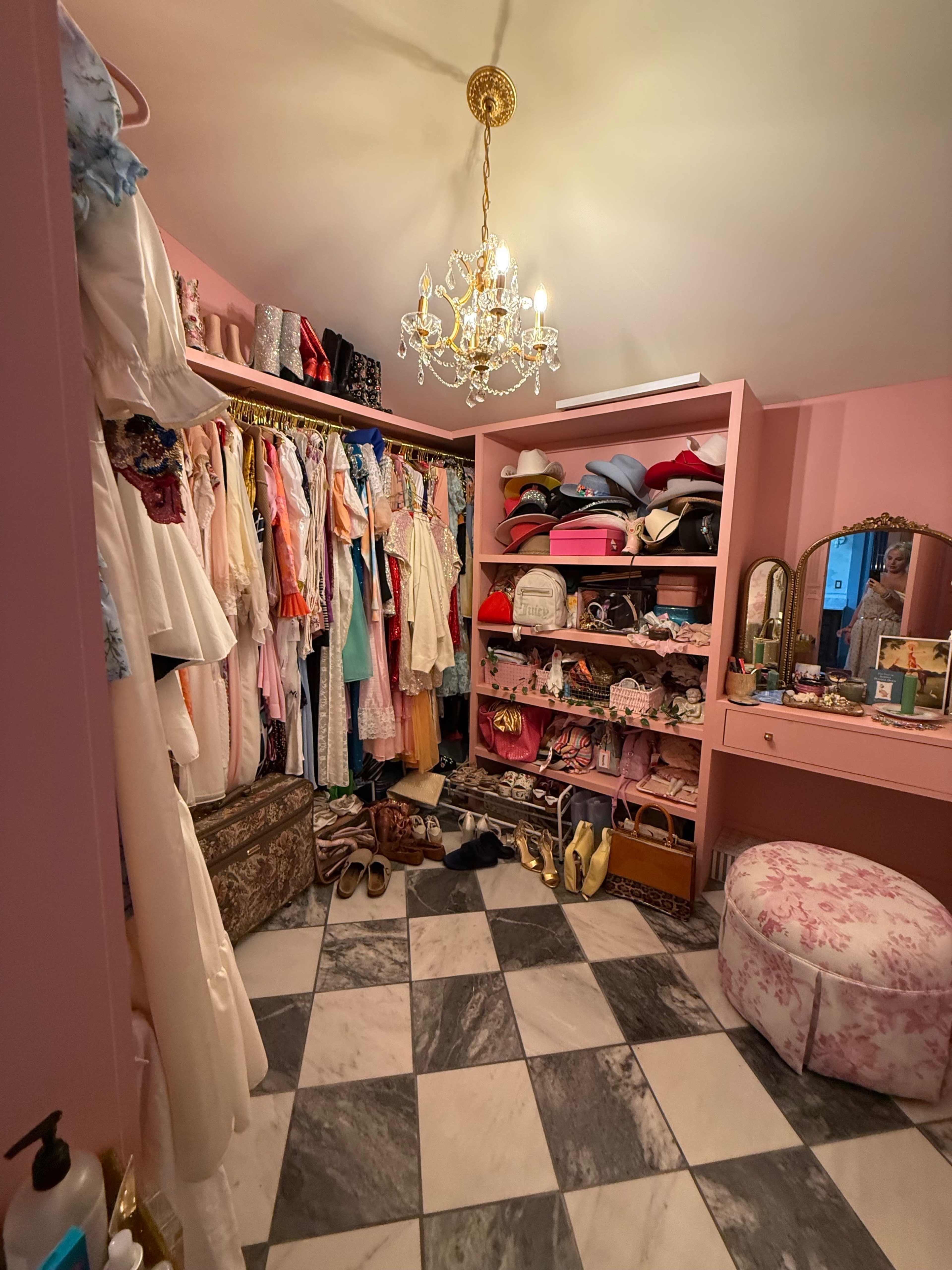 The image shows a neatly organized closet with a variety of colorful clothing, hats, and accessories arranged on shelves and hanging from rods, complemented by a chandelier and a marble-patterned floor.