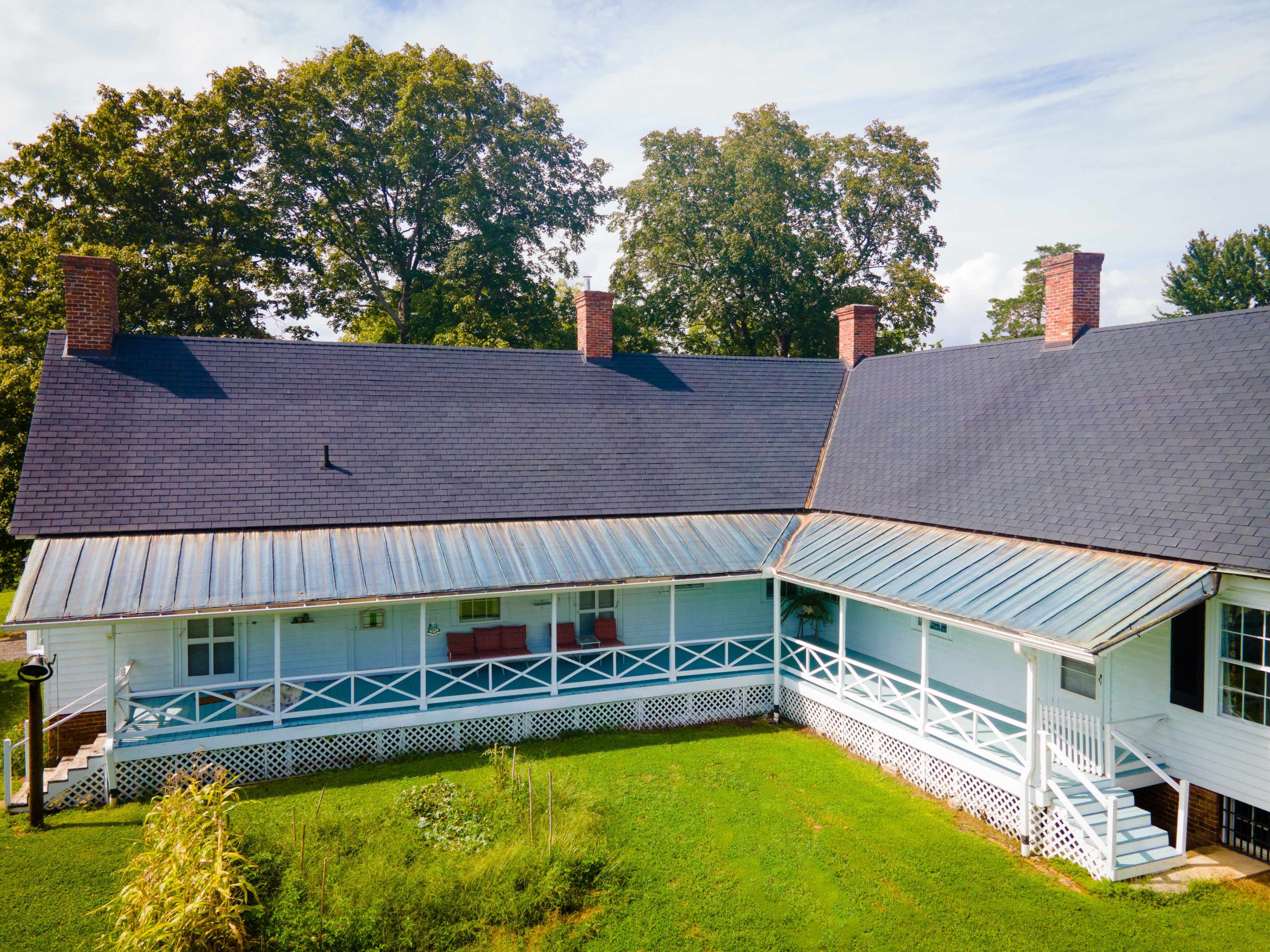 A large, two-part white farmhouse with a blue metal roof and green lawn, surrounded by trees.